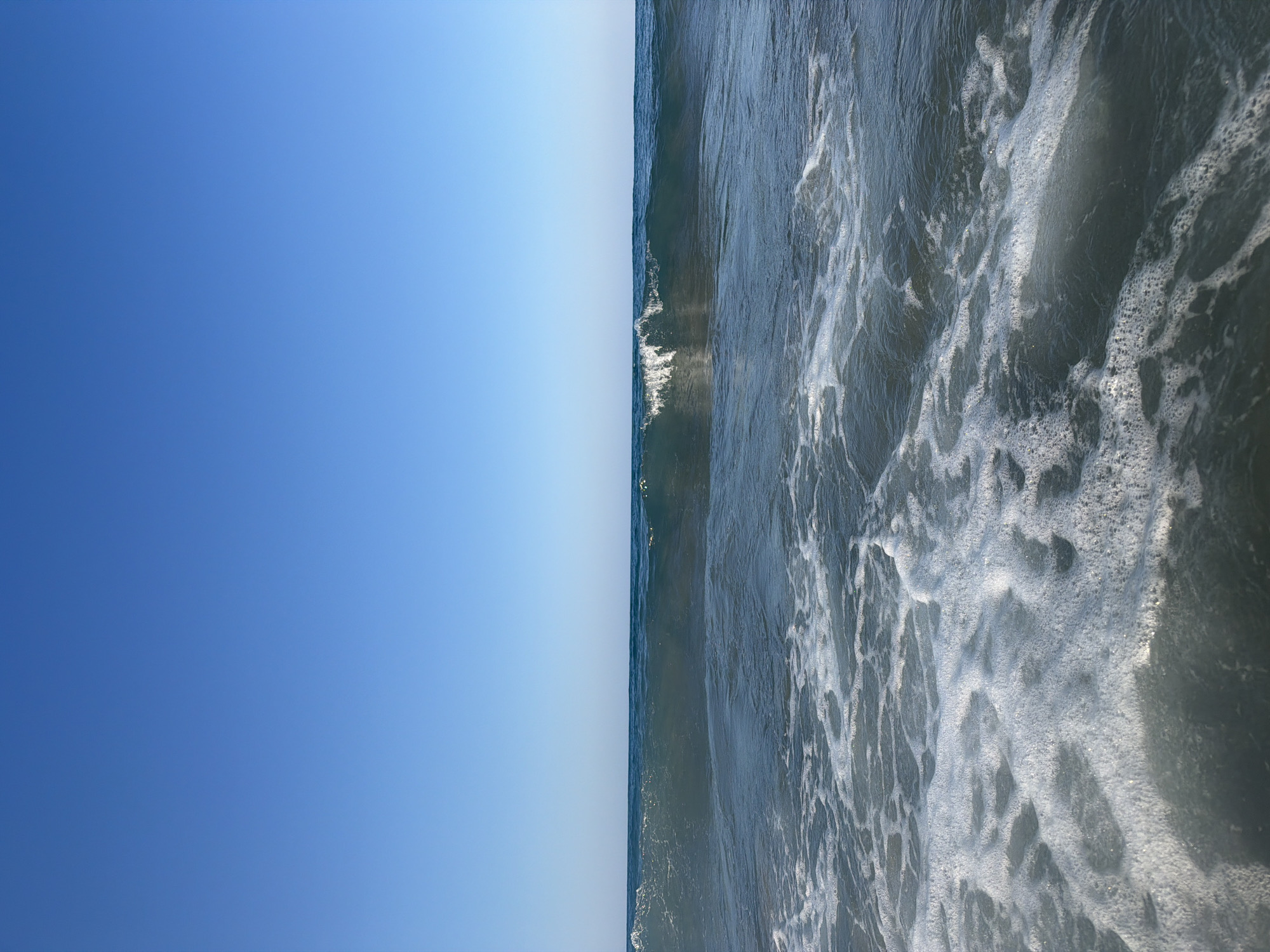 A wide, clear blue sky dominates the upper portion of the image, transitioning to the turquoise sea horizon line below. A moderate wave is breaking in the mid-ground, with white foam cresting and spilling. The foreground is filled with swirling, foamy water, suggesting the wave is washing ashore or receding. The scene depicts a natural coastal environment, likely the beach at Georgioupoli, Greece, under clear daylight conditions. There are no people or man-made objects visible in the frame.