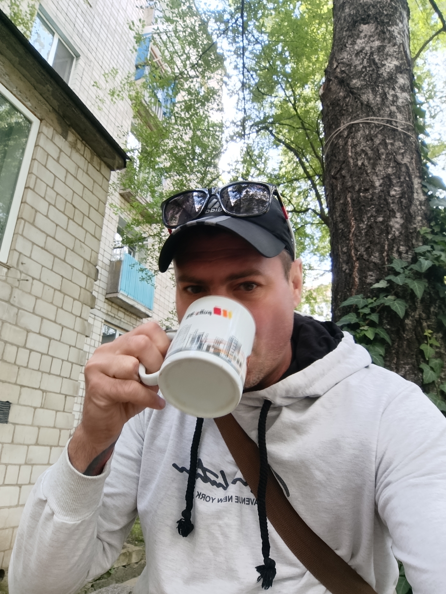 A man, wearing a light grey hoodie and a black baseball cap with sunglasses perched on top, drinks from a white mug. The mug has an illustration of buildings on it. He is standing outdoors, with a brick building to his left and a large tree to his right. The building appears to be an apartment building, with windows and balconies visible. The tree is tall and has ivy growing on its trunk. The background is filled with green foliage and a bright sky, suggesting it is daytime. The man's face is partially obscured by the mug as he takes a sip. The scene suggests a casual moment outdoors, possibly in a residential area of Ladyzhyn, Ukraine, given the architectural style and the presence of greenery typical of Ukrainian cities.