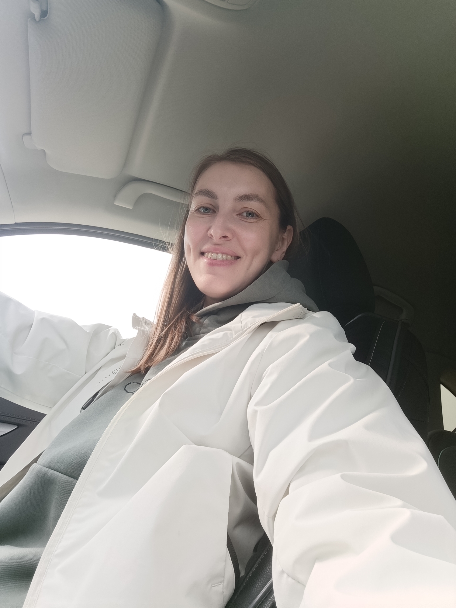 A woman is taking a selfie in the driver's seat of a car. She is wearing a light gray hoodie and a white jacket. Her hair is light brown and she is smiling. The interior of the car is visible, including the dashboard, steering wheel, and the headrest of the passenger seat. The location is likely Chaikovsky, Russia, but there is no corroborating information visible in the image to confirm.