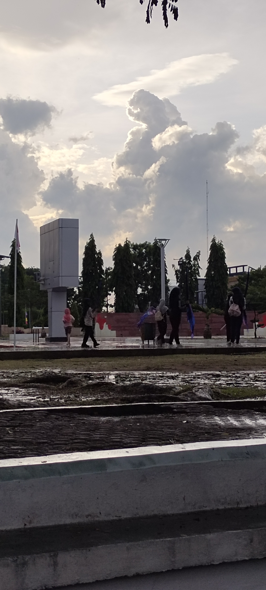 In Lhokseumawe, Indonesia, a cloudy sky looms over an outdoor area with a paved walkway and a waterlogged foreground. Several individuals, mostly obscured by distance and lighting, are visible on the walkway. One person in a pink head covering walks from left to right. Further to the right, a group of people, including those wearing hijabs, are walking or standing. One person carries a backpack. In the background, a tall, modern, rectangular structure stands among manicured trees, with a flag on a flagpole to its left. A radio tower is visible in the far right background. The ground in the foreground appears to be a cultivated field or garden bed with dark soil and some water. The overall scene suggests a public space, possibly a park or plaza, experiencing overcast weather.