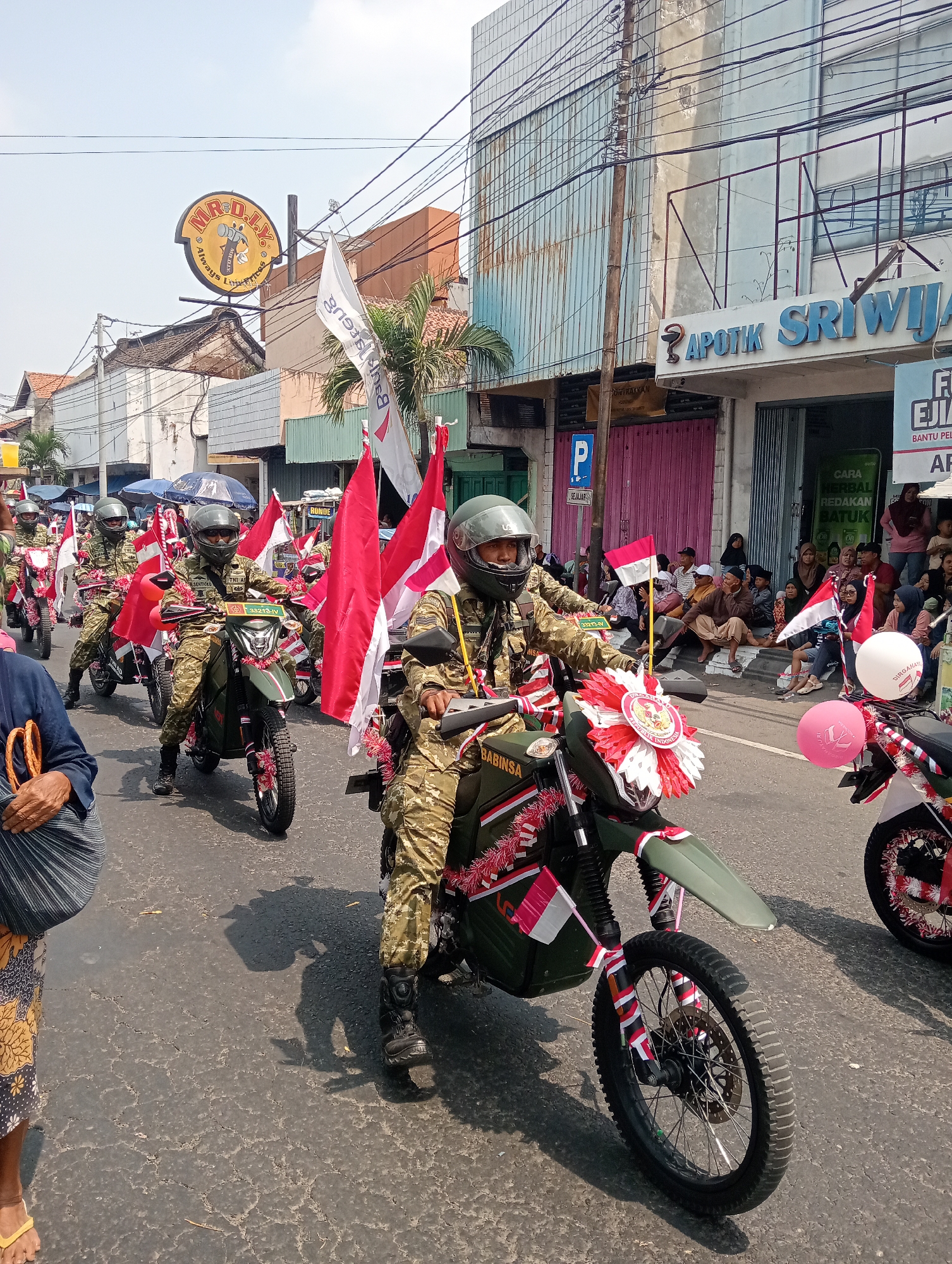 A group of men in camouflage uniforms and helmets are riding motorcycles decorated with Indonesian flags and red and white streamers in a parade. The motorcycles are green, and some are adorned with red and white pom-poms. The men appear to be part of a military or paramilitary organization, as suggested by the uniforms and the inscription "BABINSA" visible on one of the motorcycles.
The parade is taking place on a paved street in what appears to be a city. Buildings line the street, including a building with a sign that reads "APOTIK SRIWIJAYA". There are spectators on the side of the road, some sitting and watching the parade. The sky is clear and the sun is shining, indicating it is daytime.
The location is identified as the city of Demak, Indonesia, based on the presence of the "APOTIK SRIWIJAYA" sign which is a known pharmacy chain in Indonesia, and given the context of a parade with Indonesian flags. The overall scene depicts a patriotic and celebratory event.