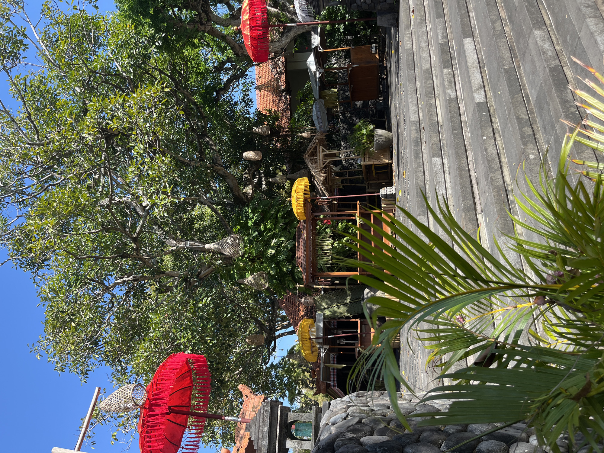This image depicts an outdoor scene in Kuta Selatan, Indonesia, likely a market or dining area. In the foreground, lush green palm fronds partially obscure a wide set of stone stairs. Behind the stairs, several market stalls with thatched roofs and wooden structures are visible, some adorned with yellow and white umbrellas. Red ceremonial umbrellas with ornate fringes are prominently displayed on the left and right sides of the image. Large trees with dense green foliage dominate the background, with woven basket-like lanterns hanging from their branches. Traditional Balinese architecture, characterized by terracotta tile roofs and stone elements, can be seen behind the market stalls. The sky is a clear, vibrant blue, indicating sunny weather. The scene appears quiet and devoid of people.