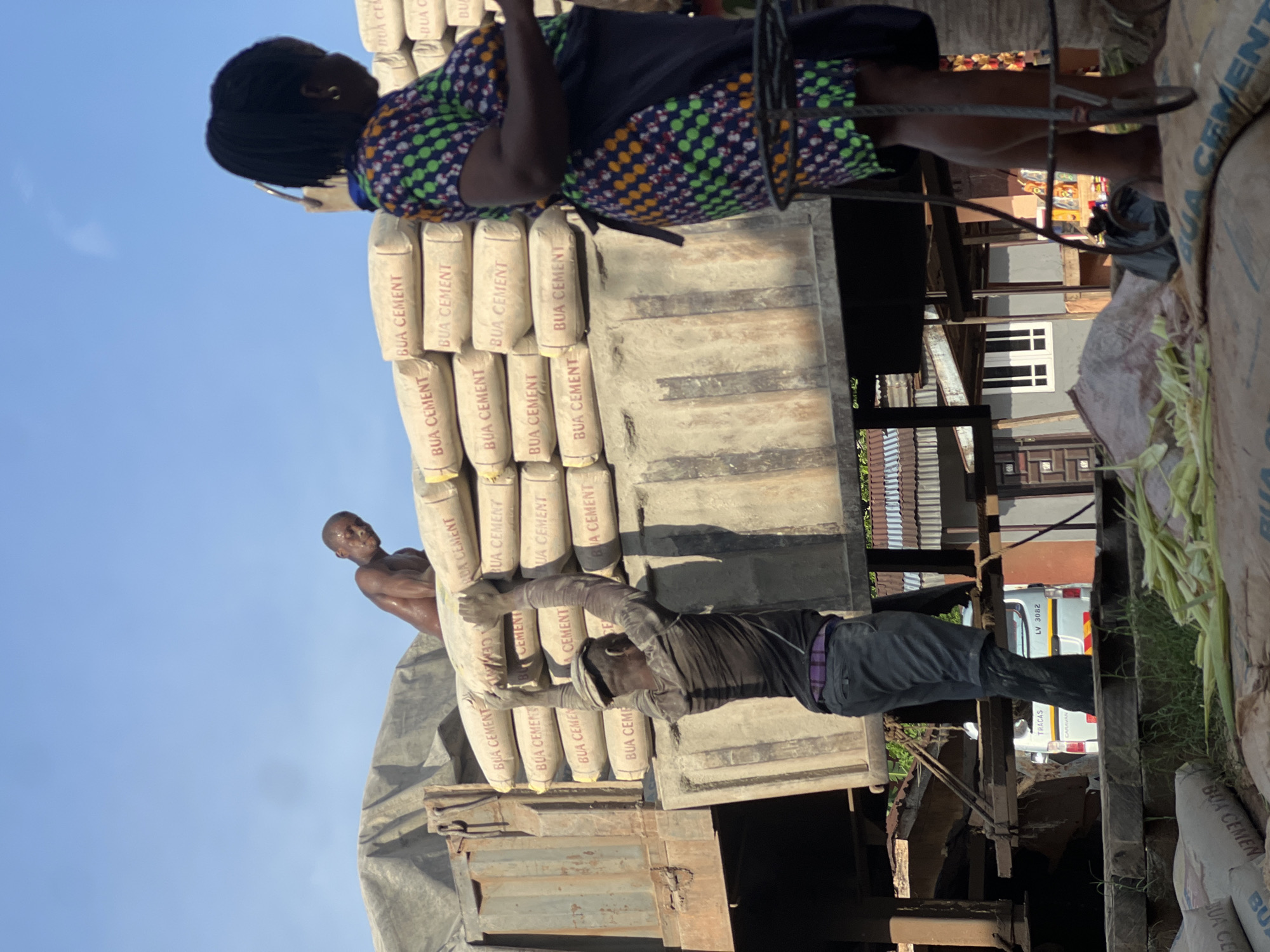 A medium shot captures a scene in Nkpor, Nigeria, with a focus on manual labor and commerce. In the foreground, several large sacks of cement, labeled "BUA CEMENT," are piled up. A man, shirtless and appearing to be sweating, is positioned on top of a truck, looking down. Another man, wearing a dark long-sleeved shirt, a cap, and dark pants with a purple belt, is actively engaged in lifting a sack of cement from the truck. His posture suggests exertion. To the right, a woman, wearing a vibrantly patterned dress and an apron, stands facing away from the camera, holding a phone to her ear. She is partially obscured by a metal structure. In the background, the truck bed is loaded with more sacks of cement, covered in part by a tarp. Beyond the truck, a street scene unfolds with buildings, including a white van with the license plate "LV 3082" parked on the side. The sky is clear blue, indicating daylight. The overall impression is one of a busy marketplace or construction site where heavy materials are being moved.