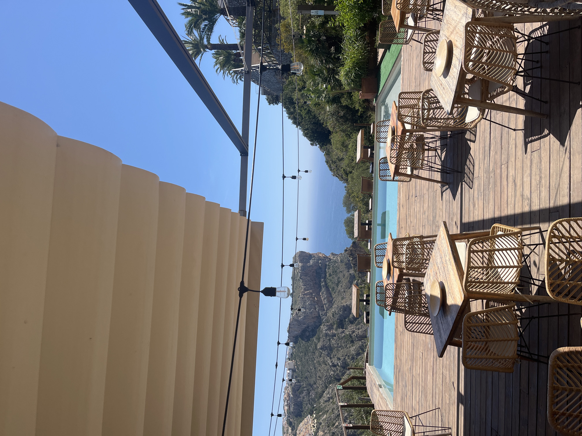The image shows an outdoor dining area in Urbanització El Cim Del Sol, Spain, overlooking a mountainous landscape and the sea. There are several wooden tables and wicker chairs set up on a wooden deck next to a long, narrow swimming pool. The tables are set with plates. String lights are strung overhead. A large, cream-colored, retractable awning is partially visible on the left side of the frame. Palm trees and other vegetation are visible in the background. The sky is clear and blue.