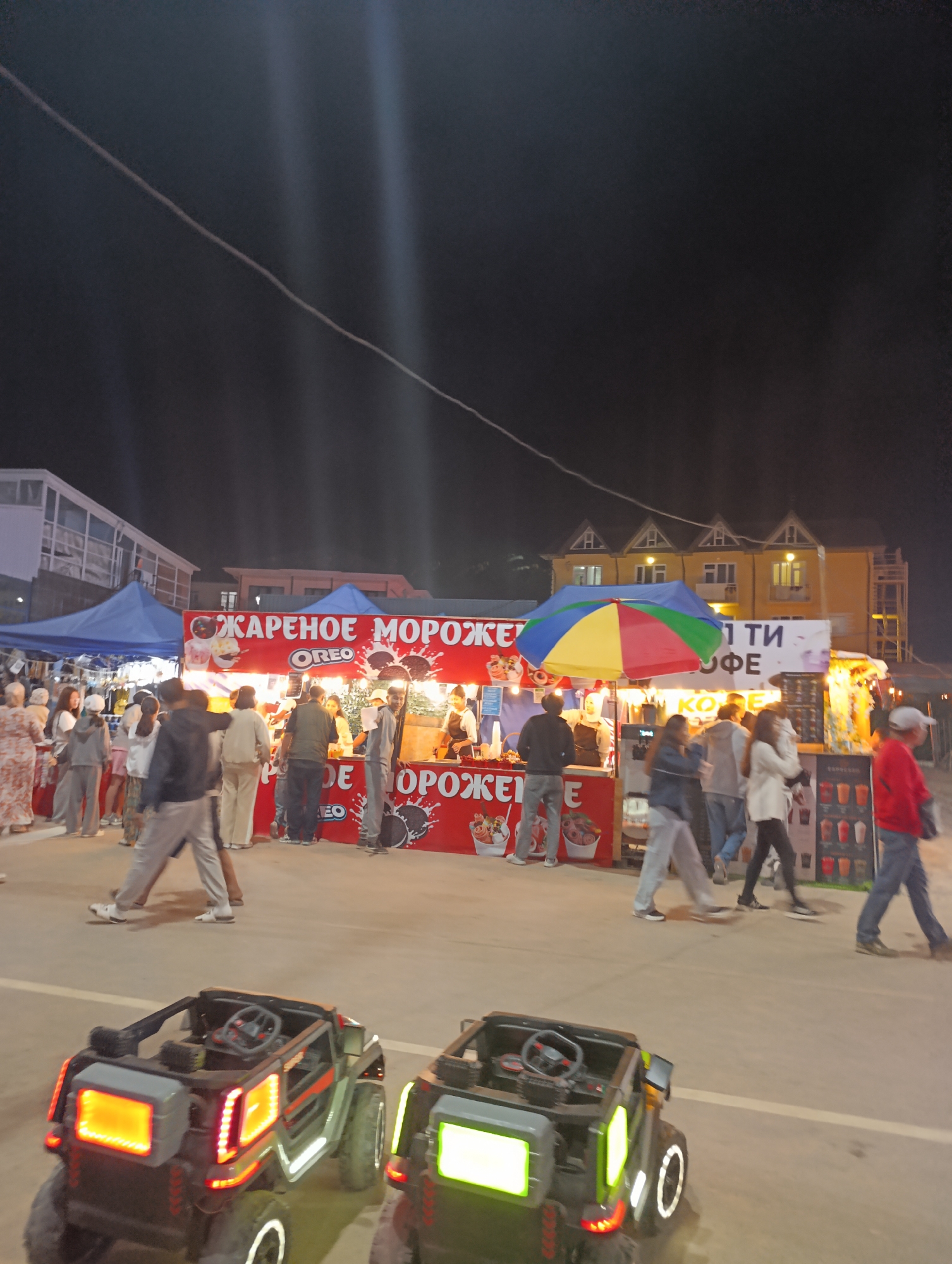 In Bosteri, Kyrgyzstan, at night, an outdoor marketplace bustles with activity. A bright red food stall with "ЖАРЕНОЕ МОРОЖЕНОЕ" (Fried Ice Cream) and "OREO" prominently displayed, serves customers. Several individuals stand at the stall, interacting with vendors. To the right, another stall advertises "КОФЕ" (Coffee) and features a colorful, multi-paneled umbrella. Numerous people are walking through the marketplace, some appearing to browse the offerings. In the foreground, two toy electric cars, designed to resemble jeeps and equipped with illuminated lights, are parked on the pavement. The background reveals buildings, including a yellow structure with distinct triangular rooflines, and tents. The scene is illuminated by artificial lights, creating a vibrant nighttime atmosphere.