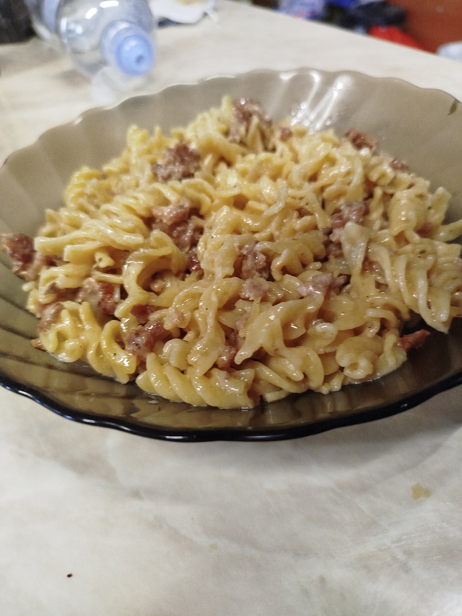 A close-up, eye-level shot shows a portion of pasta with meat, served in a dark-rimmed, wavy-edged bowl. The pasta, which appears to be rotini, is coated in a creamy sauce and mixed with small pieces of cooked meat. The background is blurred, but a water bottle is visible on the left, and the surface the bowl rests on is a light-colored, possibly marble or laminate tabletop. The image is taken indoors, likely in a home setting. There are no people or animals present, and no specific location within Zavodoukovsk, Russia is identifiable.