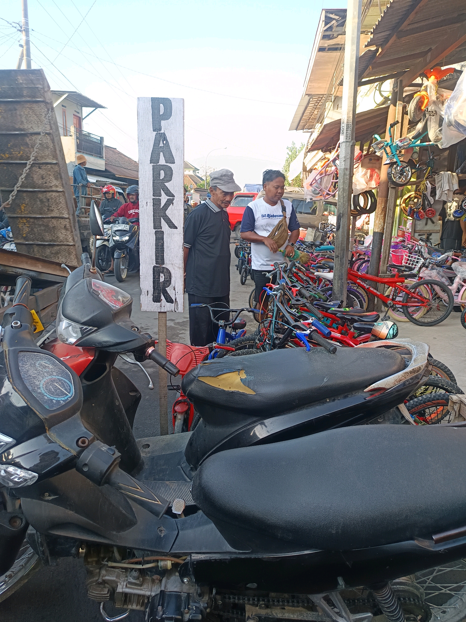 The image shows a street scene in Blega, Indonesia, likely a market area, with a variety of vehicles and stalls. In the foreground, two motorcycles are parked, one black and the other red. A tall, white sign with the word "PARKIR" (parking in Indonesian) is prominently displayed.
To the right of the sign, two men are interacting. One man, wearing a black shirt and a grey cap, appears to be looking at the other man, who is wearing a white t-shirt with blue trim and carrying a brown bag over his shoulder. Behind them, a stall is filled with colorful bicycles of various sizes. Further back, more motorcycles and a red car are visible.
The environment appears to be outdoors, with a clear sky overhead and a corrugated metal roof over the bicycle stall. The overall impression is of a bustling commercial area where parking and goods are being offered.