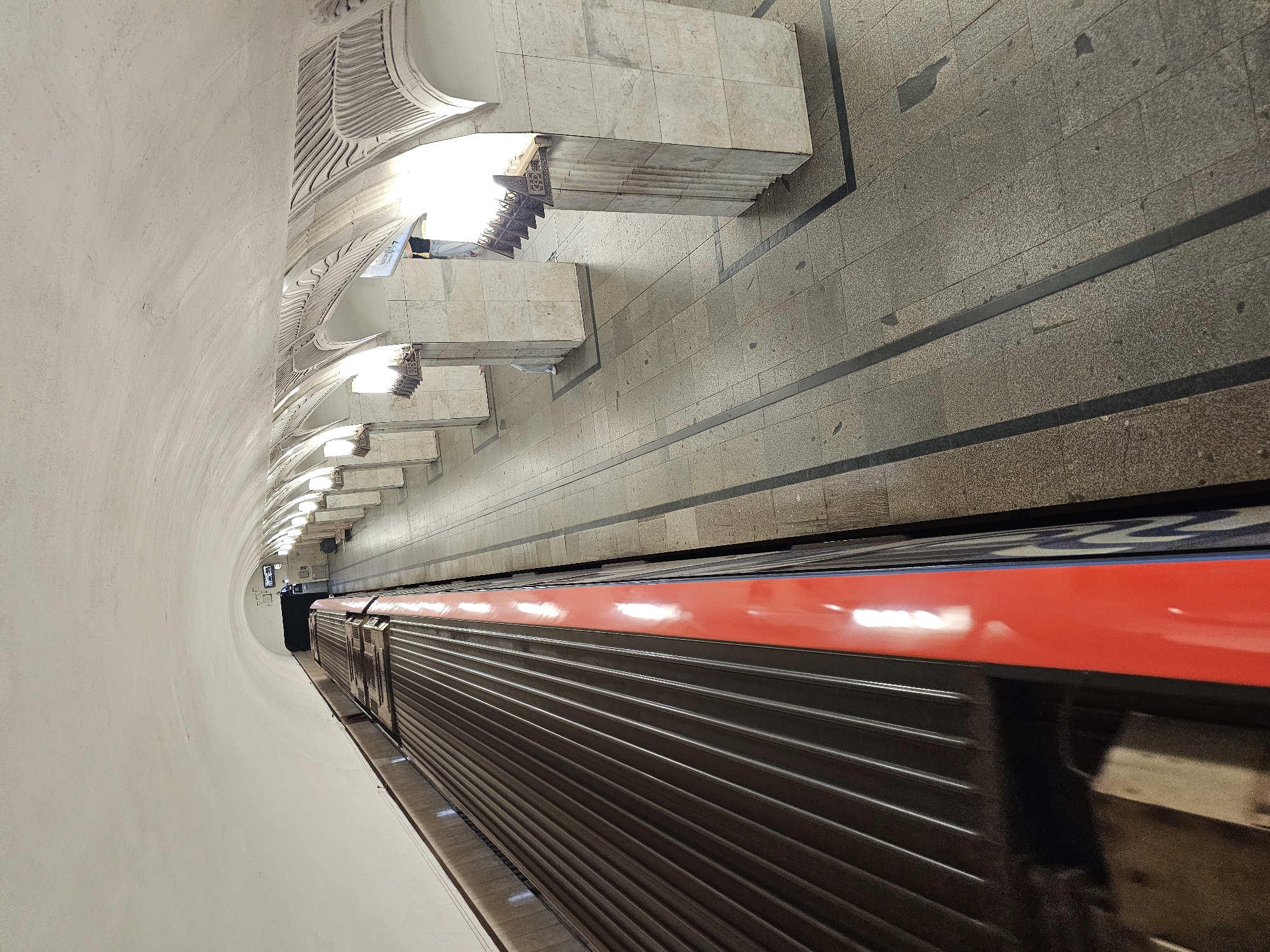 The image depicts the platform of the Moscow Metro station, likely Komsomolskaya or a similar ornate station, featuring the characteristic barrel vault ceiling and decorative arches supported by marble columns. A red and grey train is stopped at the platform, its roof visible in the foreground. The platform is paved with granite tiles and marked with black lines. Lighting fixtures are integrated into the arched structures. A few individuals are visible on the platform, one person standing near the arches and another partially visible in the distance. The station appears to be clean and well-maintained.