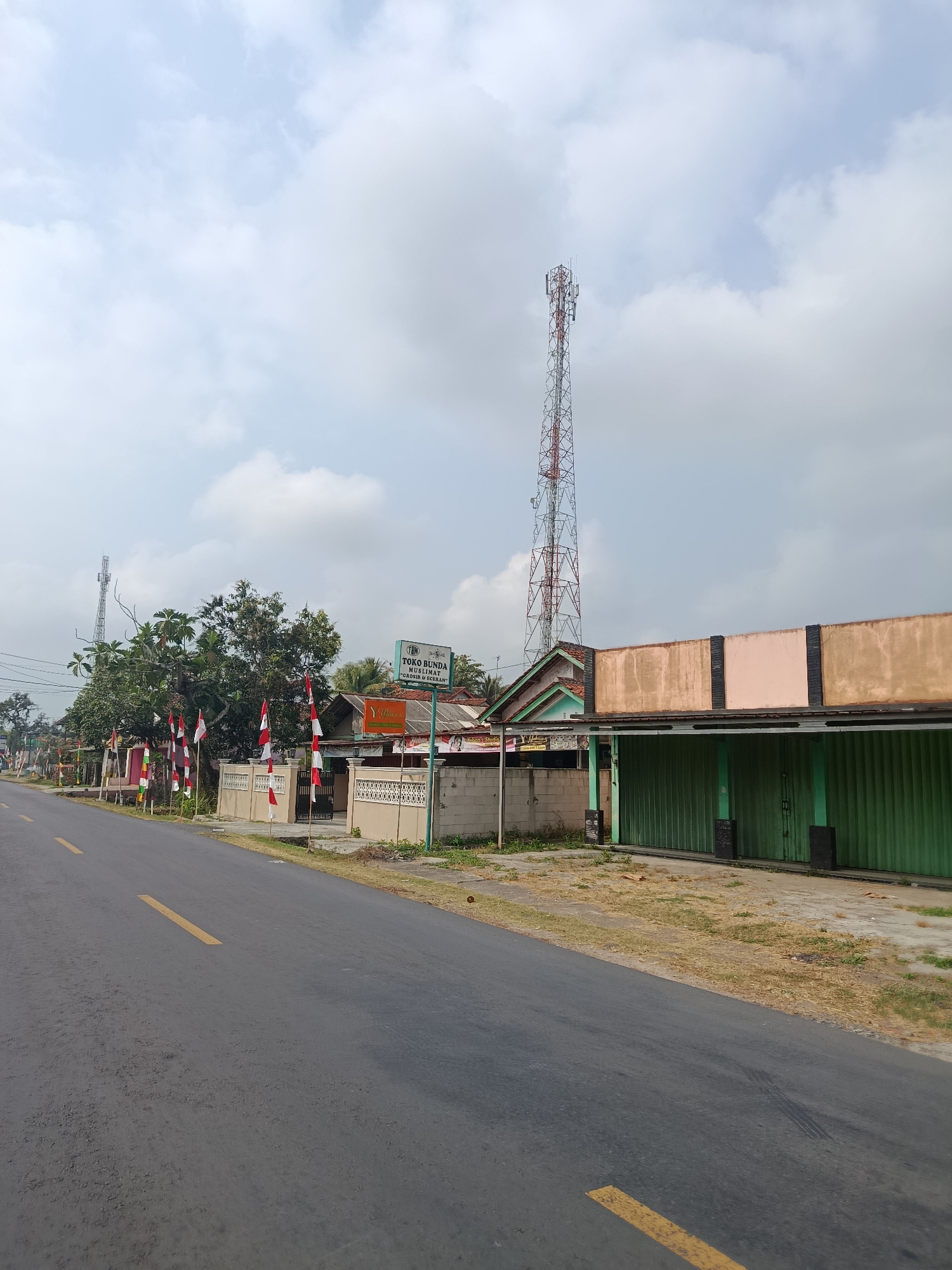 The image depicts a street scene in Bantarsari, Indonesia, under a cloudy sky. A paved road with a yellow dividing line runs diagonally from the lower left to the middle right of the frame. Along the left side of the road, a row of Indonesian flags, red and white, are planted along a low, decorative wall. Behind the flags are trees and several buildings. A prominent tall telecommunications tower with a red and white lattice structure stands in the background, to the left of a cluster of trees. To the right of the road, a long, single-story building with a green roller-door facade and an extended roofline is visible. A sign for "TOKO BUNDA MUSLIMAT" is mounted on a pole in front of this building. The environment appears to be a somewhat rural or suburban area with a mix of residential and commercial structures, and infrastructure elements like the cell tower.
