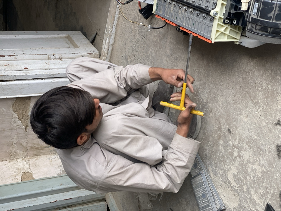 In Kandahar, Afghanistan, a young male individual, dressed in a light gray shalwar kameez, is observed squatting on a rough concrete surface. He is actively engaged in repairing or disassembling a large electronic component, identified as a multi-cell battery pack, situated to his right on the ground. He uses both hands to manipulate a yellow T-handle wrench, applying torque to a fastener on the battery pack. To his left, several metallic plates or covers are stacked on the ground. The background reveals a dilapidated wall with peeling light-colored paint and a slightly ajar white wooden door, also exhibiting significant paint loss and wear.
