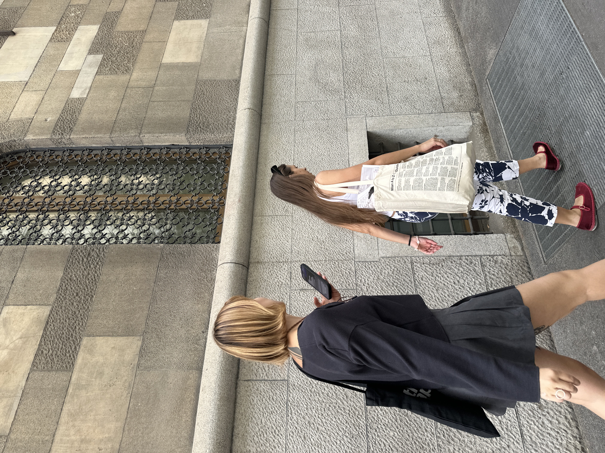 The image captures two young women walking on a city street in Milan, Italy. The woman in the foreground is holding a smartphone, wearing a short, dark-colored skirt, a black jacket, and a tote bag. Her hair is short, blonde, with dark roots. The woman behind her has long brown hair and is wearing a white tank top, floral print trousers, and red shoes. She is carrying a tote bag with the words "Milan A to Z Guide" printed on it. They are walking on a grey stone sidewalk next to a building with a decorative window grill. The street has a metallic grate on one side.