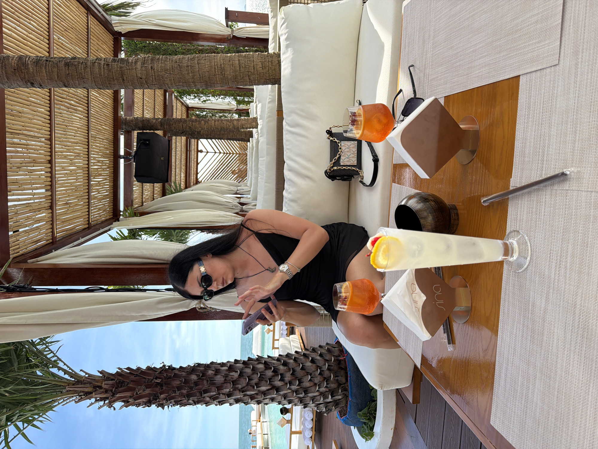 The image captures a woman seated in an outdoor lounge area in Pattaya City, Thailand. The woman is dressed in a black dress and sunglasses, and she is looking at her phone. On the table in front of her, there are two cocktails, one orange and one with a lemon garnish, along with a black purse and napkins. The lounge area is decorated with white cushions and a canopy roof, and palm trees are visible in the background. The sky and sea are also visible in the distance, suggesting a coastal location.