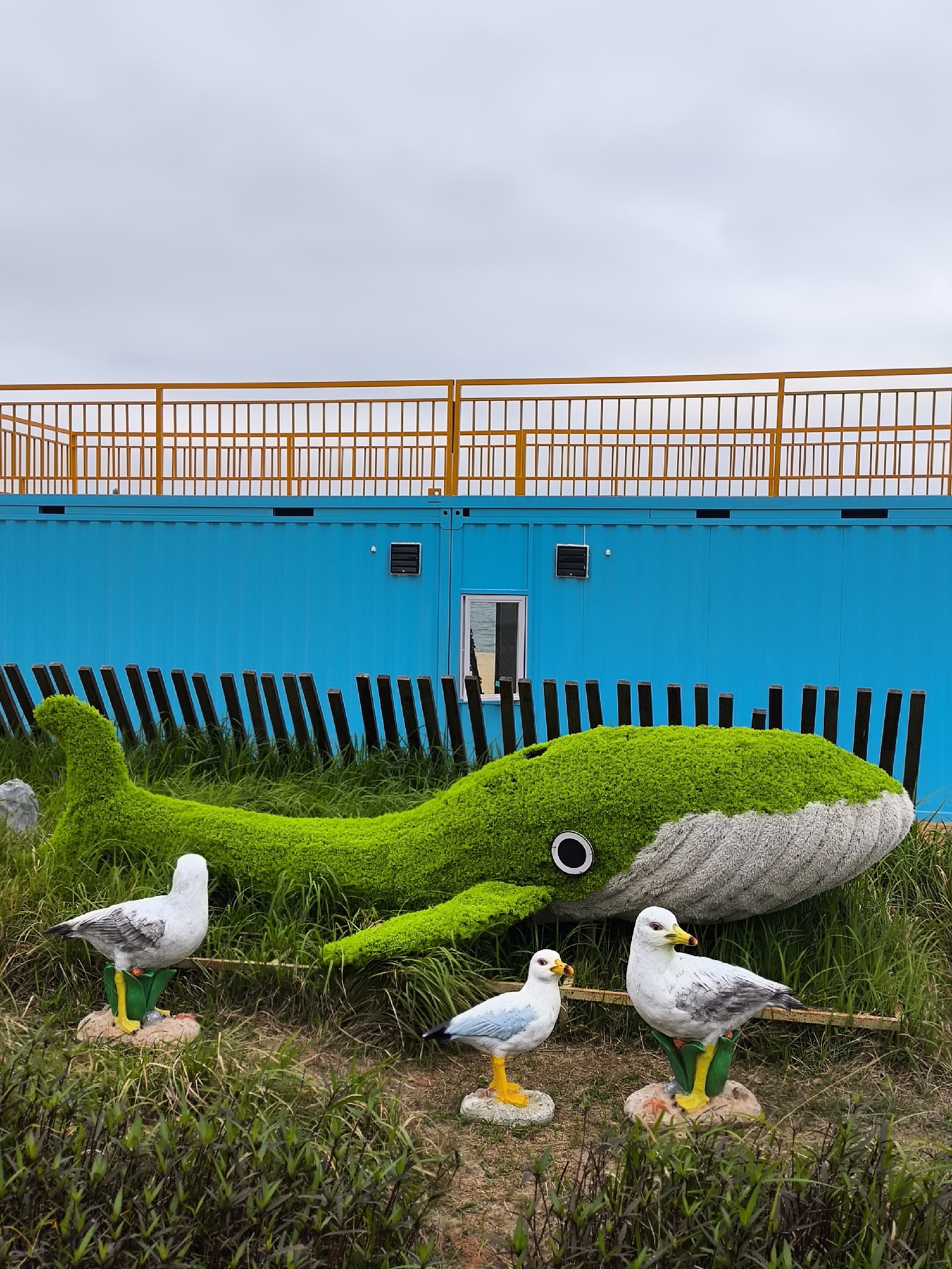 The image depicts a marine-themed exhibit outdoors in Yantian District, China. A topiary whale, covered in green moss on its head and body and a grey, gravel-like material on its tail, rests in a grassy area in front of a blue building with a yellow fence on its roof. Three statue seagulls, with white and grey plumage and yellow beaks and feet, stand on small rock-like pedestals in front of the whale. The sky is overcast. Vertical black bars are positioned behind the whale.