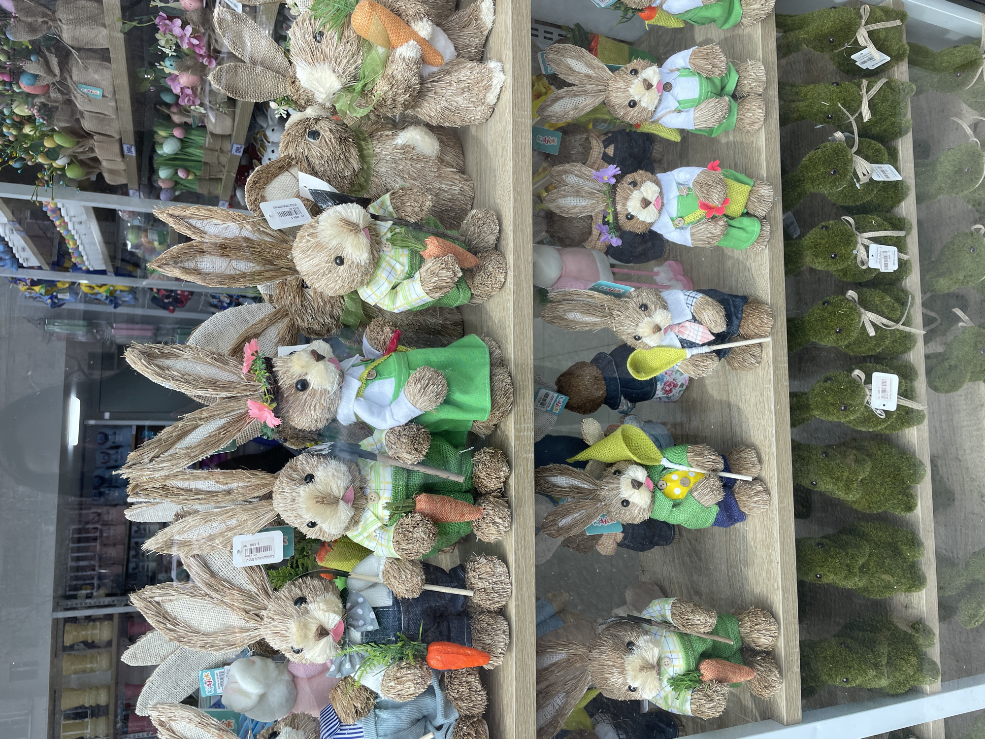 The image depicts a retail display of handcrafted Easter-themed decorations, likely within a shop window. The location is suggested to be Yerevan, Armenia.
On the left side of the image are straw bunnies with long ears and dressed in various outfits including overalls, dresses, and hats. Some hold small carrot props. The display appears tightly packed with bunnies, creating a dense grouping.
To the right of the bunnies are smaller, plush teddy bears dressed in similar spring outfits and accessorized with props like miniature shovels, flowers, and carrots.
On the far right are small, moss-covered rabbit figures with tied ribbons and price tags attached. These figures are arranged in a row.
The reflections on the glass window show the store's interior, which appears to contain additional products and shelving. The background is blurred, but it seems to show a retail environment.
