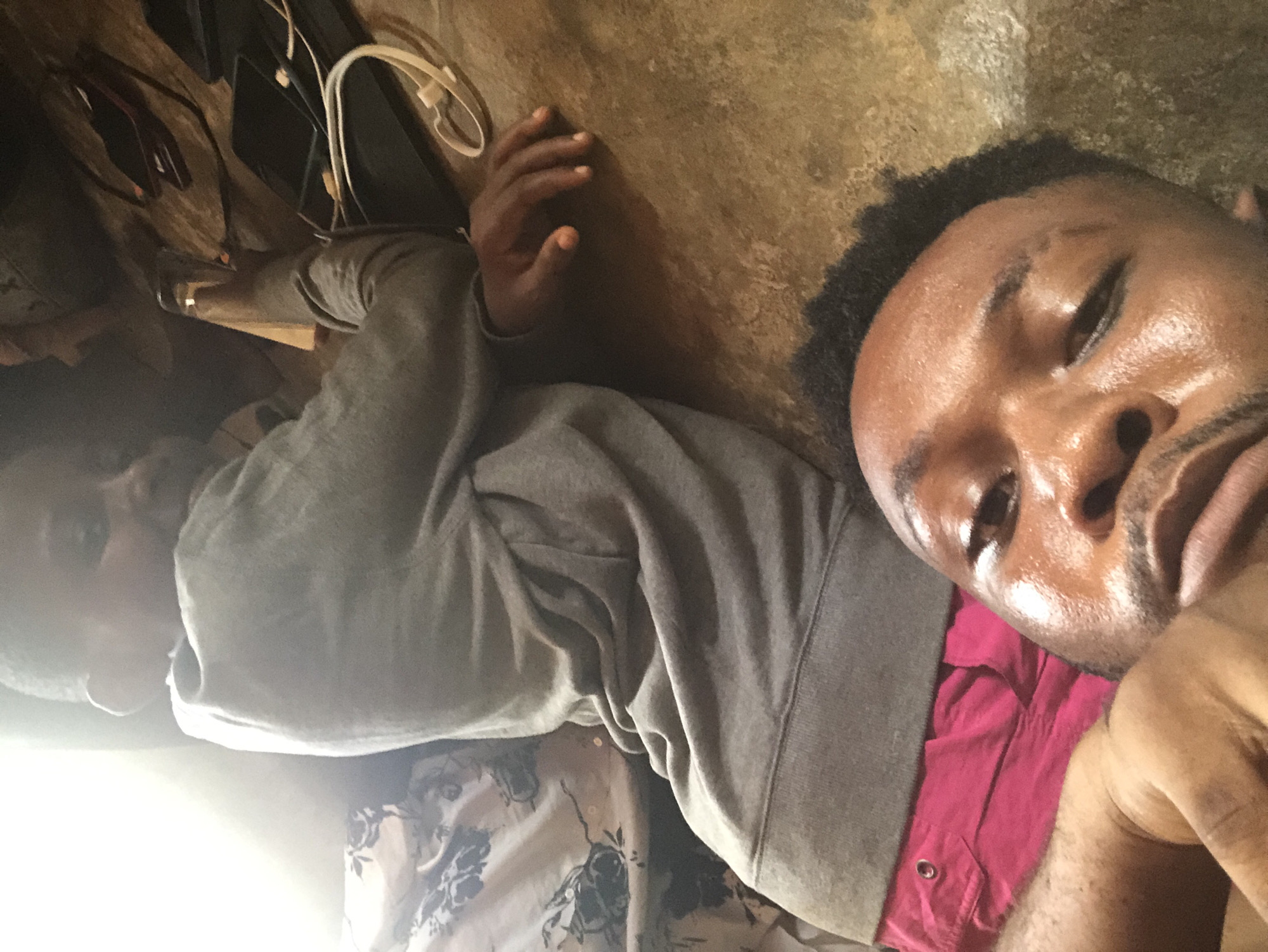 A selfie-style shot shows two individuals lying on what appears to be a concrete or tile floor. The person in the foreground is close to the camera, displaying a face with a slightly oily sheen. They are wearing a red shirt peeking out from beneath a gray sweatshirt. The second person is partially visible behind, wearing a patterned shirt and a gray sweatshirt. Cords and electronic devices are visible near the top left corner of the image. Location appears to be indoors and undetermined as the city and location are both "Unknown".
