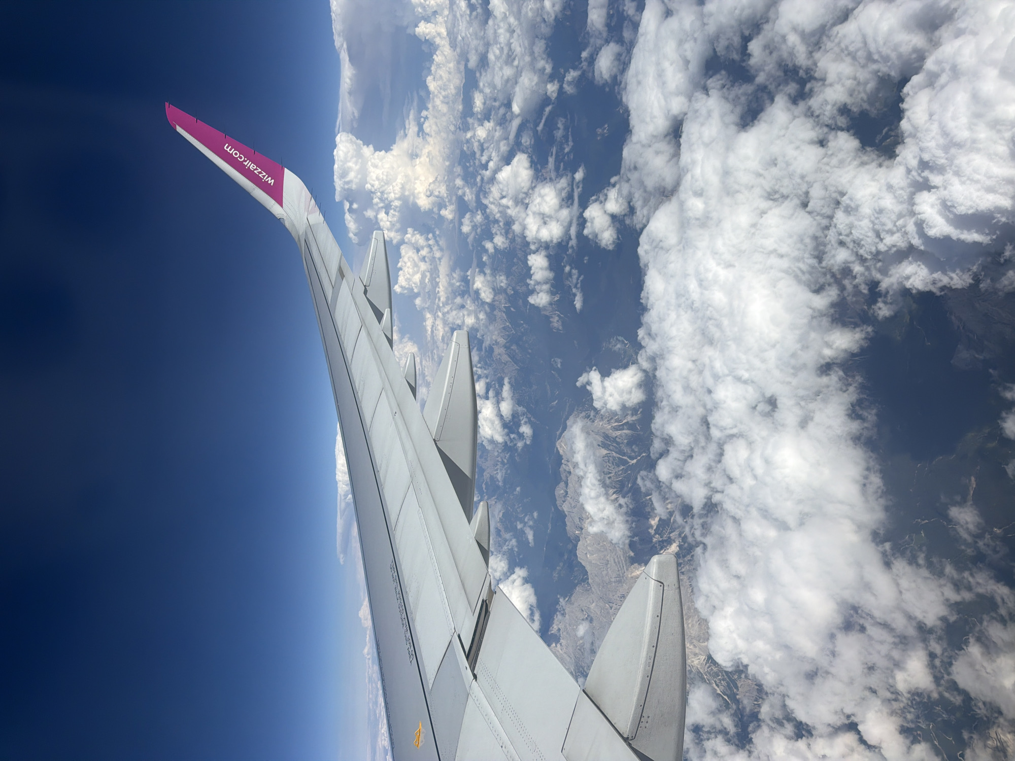 The image, taken from the window of an airplane, shows a view of the earth below and the sky above. The airplane's wing, a part of the Wizz Air aircraft, is prominently visible in the foreground, extending across the frame. The wing is white with a vibrant pink tip, featuring the "wizzair.com" logo.
Below the wing, the landscape is a mix of rugged mountains, appearing grey and sparsely vegetated, and a dense covering of white, fluffy clouds. The clouds create a textured layer, partially obscuring the terrain beneath. Glimpses of dark, possibly shadowed areas, can be seen through gaps in the clouds, suggesting valleys or darker landforms. The sky above is a deep, clear blue.
The scene depicts an aerial view, likely during daylight hours, with sunlight illuminating the clouds and the top of the wing. There is no indication of the city of Leno, Italy, or any specific identifiable landmarks within this aerial perspective. The image focuses on the airplane's wing and the natural environment seen from altitude.