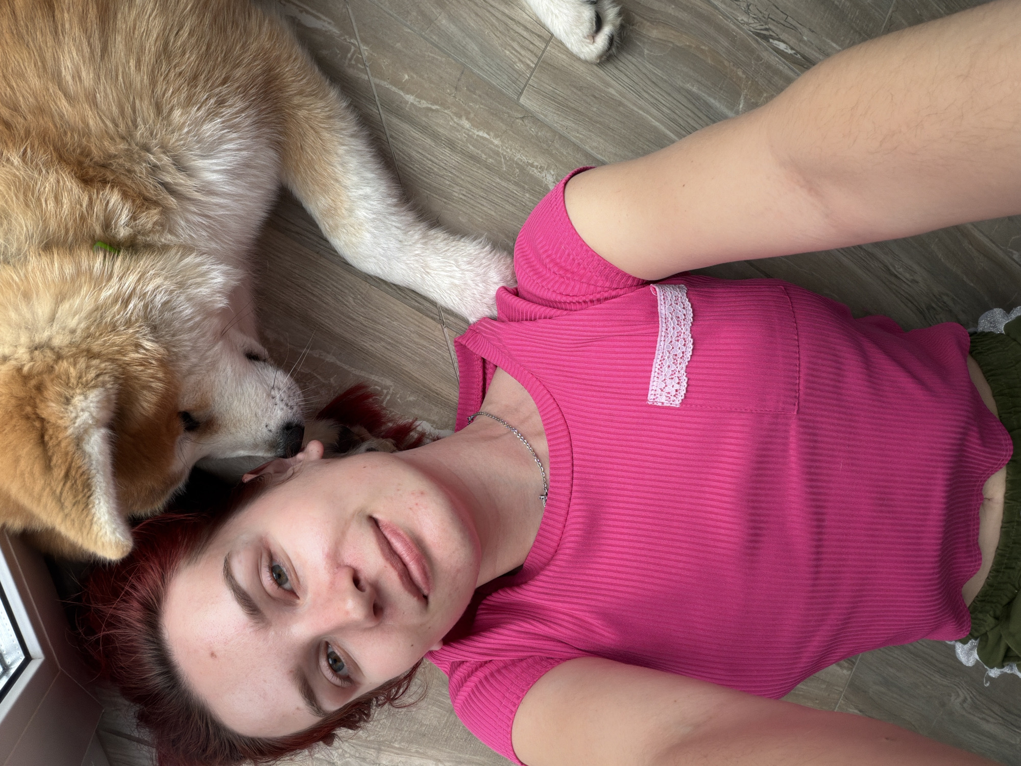A young woman and an Akita-type dog are captured lying together on a light wooden floor in an indoor setting. The woman has reddish-brown hair and green eyes, and is wearing a ribbed, fuchsia-colored short-sleeved top adorned with a small white lace detail on the left shoulder area. She also wears a silver chain necklace with a cross pendant. Lying on her back, she gazes directly at the camera with a relaxed, slightly smiling expression. The dog, predominantly tan and white-furred, rests its head close to the woman's, with only a portion of its body and one paw visible. A window frame is partially visible in the upper left background, suggesting natural light during the daytime. The scene conveys a sense of calm and affectionate companionship between the owner and her pet. No text is visible in the image.