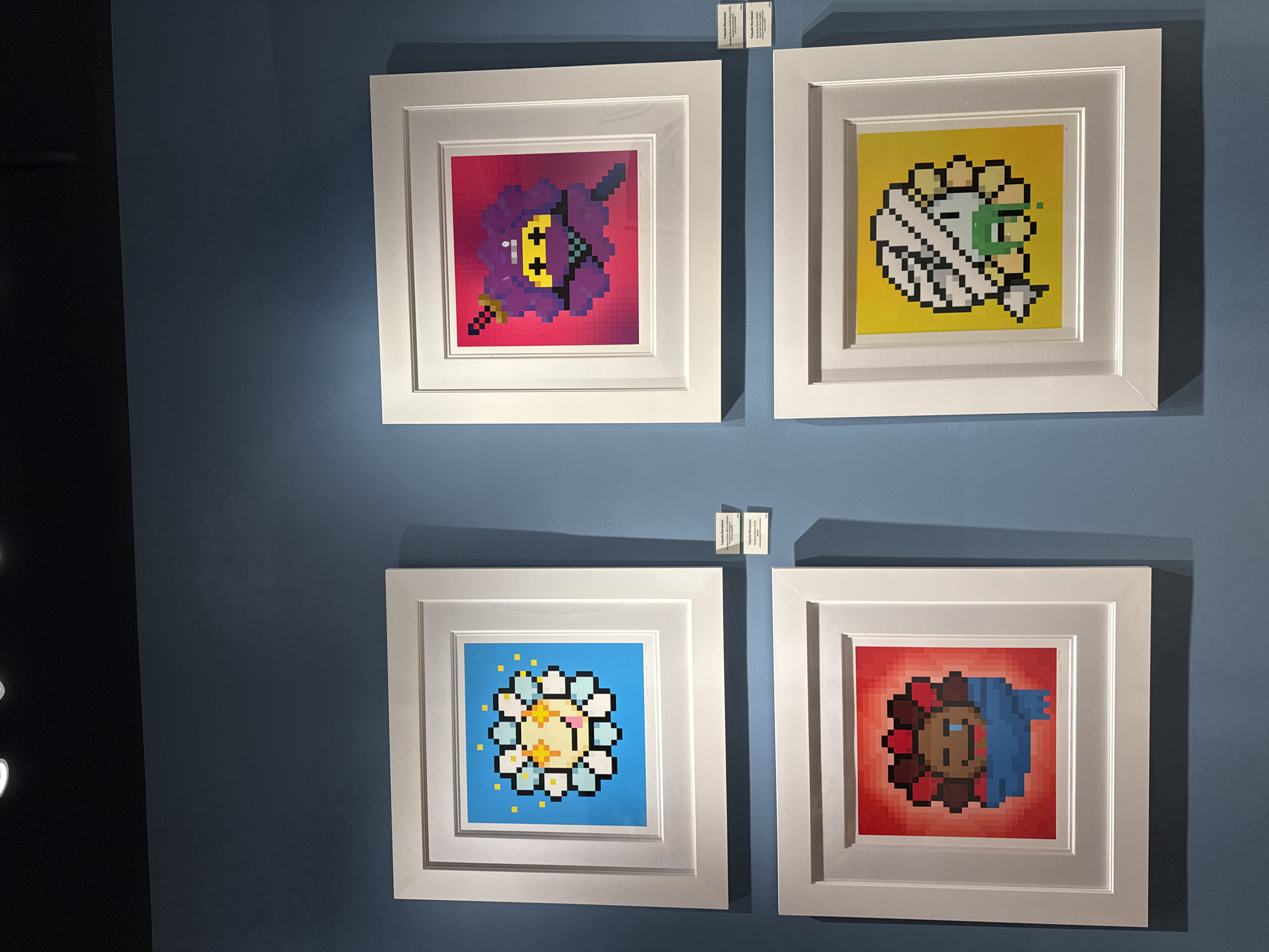 Four framed, pixelated, floral-themed artworks hang on a blue wall. The frames are white. The top left artwork features a purple pixelated flower with a yellow face against a pink background. The top right artwork is a white pixelated flower with a green center against a yellow background. The bottom left artwork displays a yellow and white pixelated flower against a blue background. The bottom right artwork shows a brown and blue pixelated flower against a red background. Small labels are visible above the top two frames. The scene likely occurs indoors, given the wall and lighting. I cannot identify the specific location in Schaumburg, United States of America, from this image alone.