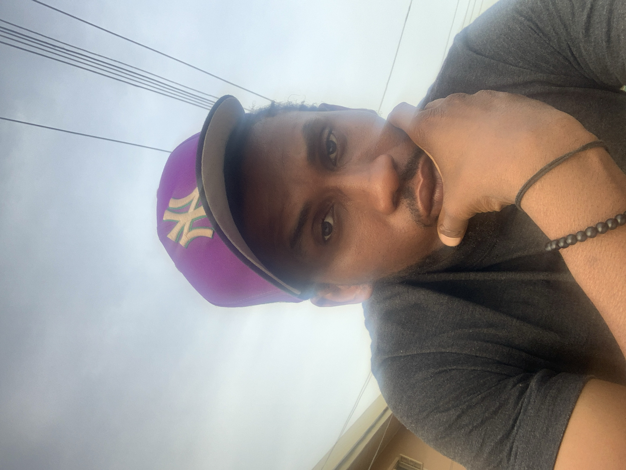 A man is depicted in a close-up, slightly tilted shot. He is wearing a purple baseball cap with a gold logo, and a dark grey or black t-shirt. He has dark skin and facial hair. His right hand is raised near his face, with his fingers touching his cheek. He is wearing two bracelets on his right wrist. The background is a bright, overcast sky with power lines visible. A building with tan walls is partially visible in the lower right corner of the frame. There is no information to determine the location as Jos, Nigeria.