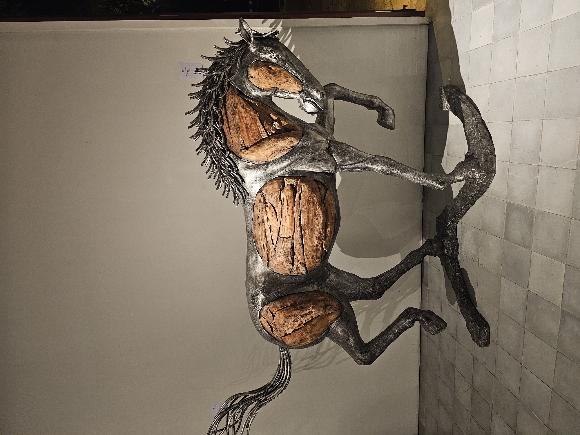 The image shows a metal and wood sculpture of a rearing horse. The outer body of the horse is constructed from metal, while the interior portions of the neck, torso, and rump are filled with what appears to be rough-hewn wood blocks. The horse's mane and tail are composed of thin metal rods. The sculpture is positioned against a wall that appears to be divided, with one side being a plain beige surface and the other side tiled with a grid of square tiles. The image was taken in Denpasar, Indonesia.