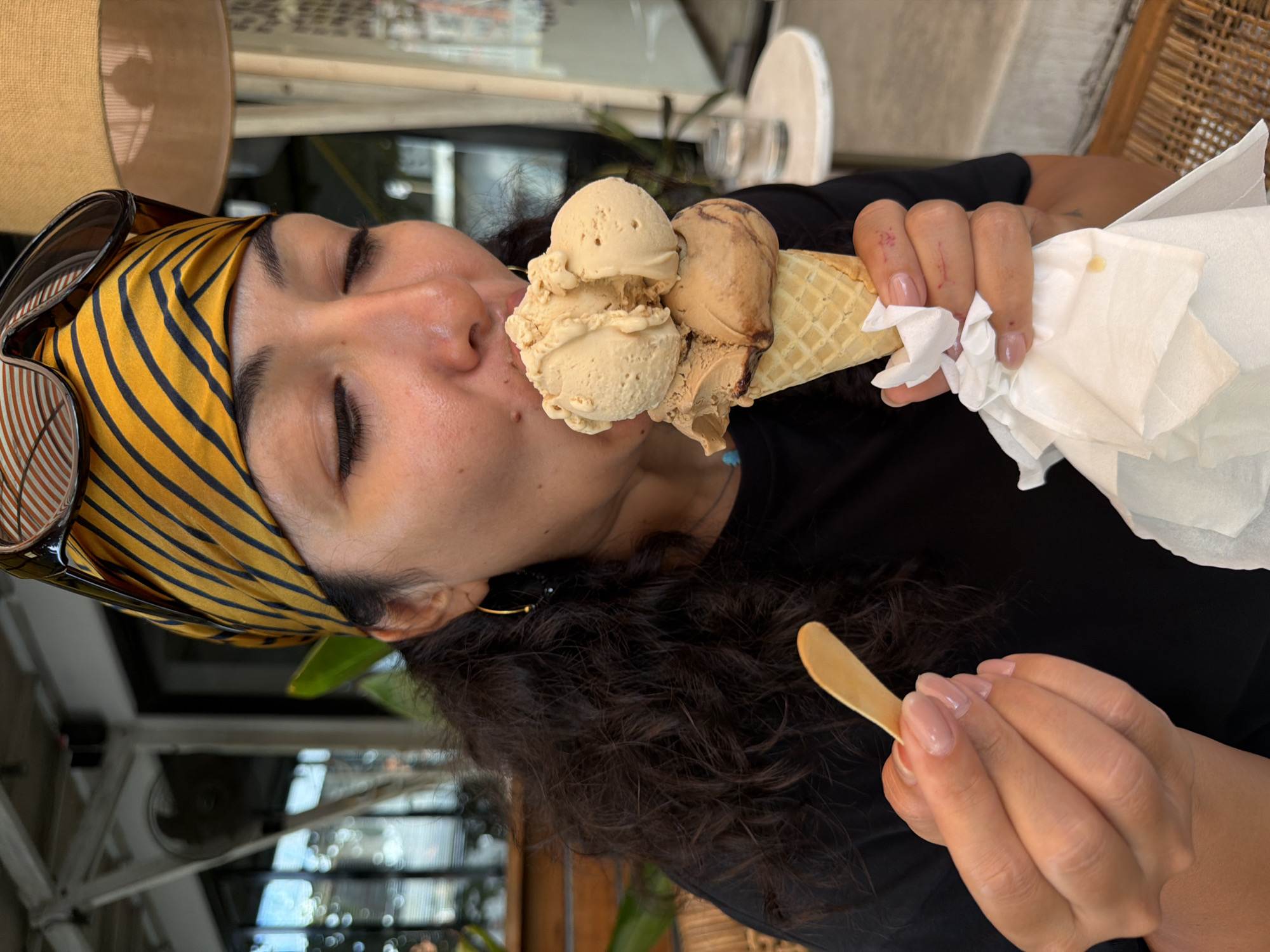 The image shows a woman indoors, likely in a café or similar establishment in North Kuta, Indonesia, as implied by the setting and background elements. She is wearing a yellow and black striped headscarf and sunglasses perched on top of her head. The woman is in the act of eating an ice cream cone with at least three scoops of light brown and tan ice cream. In her other hand, she holds a small wooden spoon. A wad of white napkins is wrapped around the base of the cone. She has dark curly hair and is wearing a black top. The background includes elements of the establishment such as a lamp with a beige shade, a white table, and various reflective surfaces suggesting glass or mirrored elements.