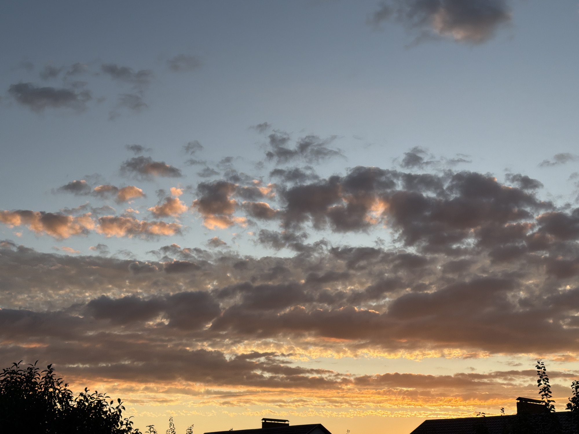The image captures a sunset sky over the city of Uzyukovo, Russia. The sky is filled with a variety of clouds, ranging from dark, heavy formations to lighter, wispy clouds tinged with orange and pink from the setting sun. The lower portion of the image shows the silhouettes of rooftops and trees, indicating a residential or semi-urban environment. The light suggests it is either early morning or late evening.