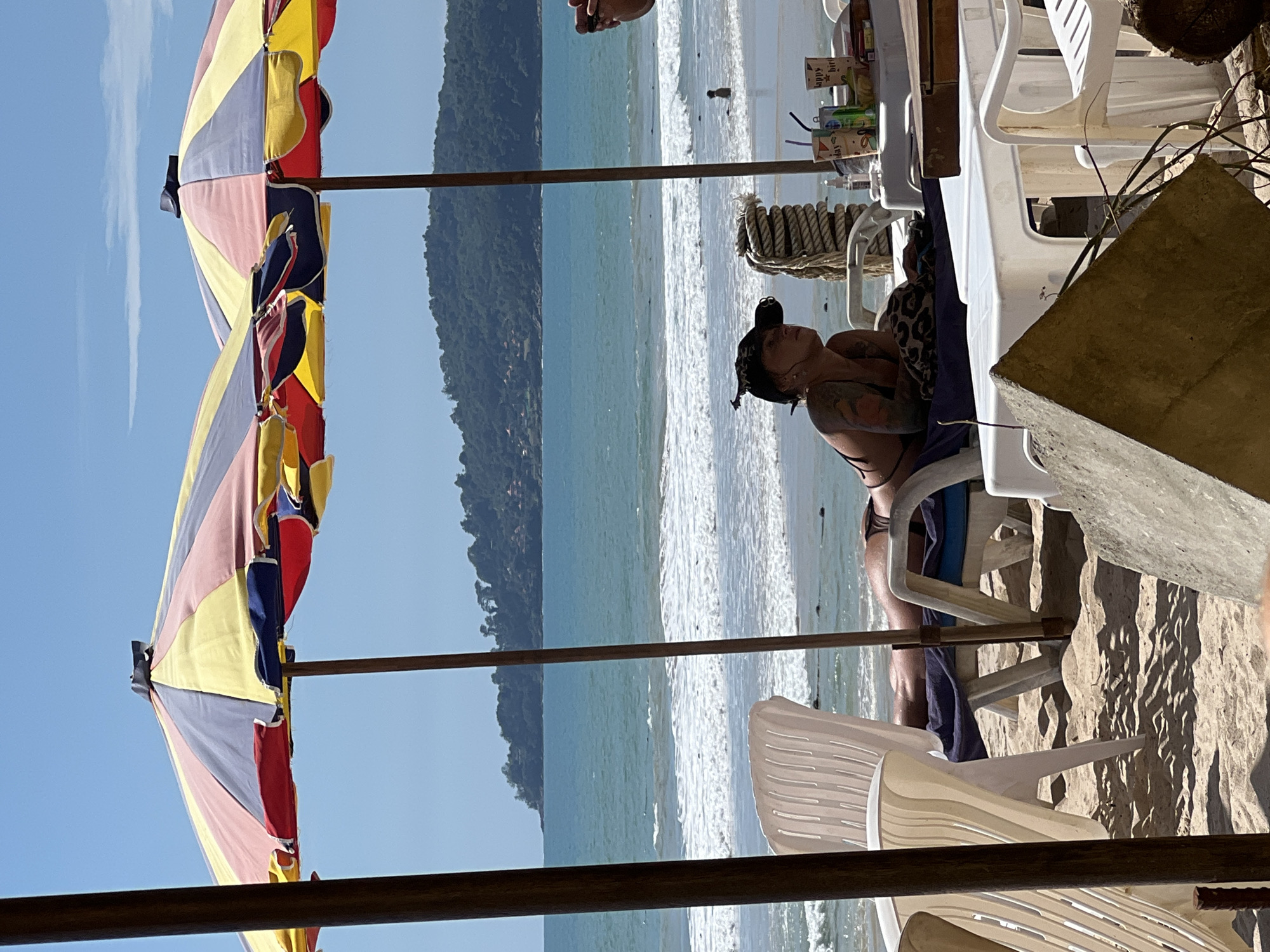 The image depicts a beach scene in a location of Unknown, Unknown, under a clear blue sky. In the foreground, two multi-colored beach umbrellas, one predominantly yellow and red, and the other with sections of yellow, blue, pink, and red, are set up on the sand. A person wearing a black bikini and a black baseball cap is lying on a white lounge chair under the umbrella, facing towards the sea. The person has tattoos on their arms. To the left of the lounge chair, a white plastic chair is visible. On a table behind the lounge chair, several drinks and cups are arranged, including one with "happy birthday" printed on it. The sea, with moderate waves and white foam, stretches out to the horizon, where a lush green, tree-covered hill is visible. A small figure of a person can be seen in the water in the mid-ground. The sand is light brown and textured, with visible shadows cast by the umbrellas and foreground objects.