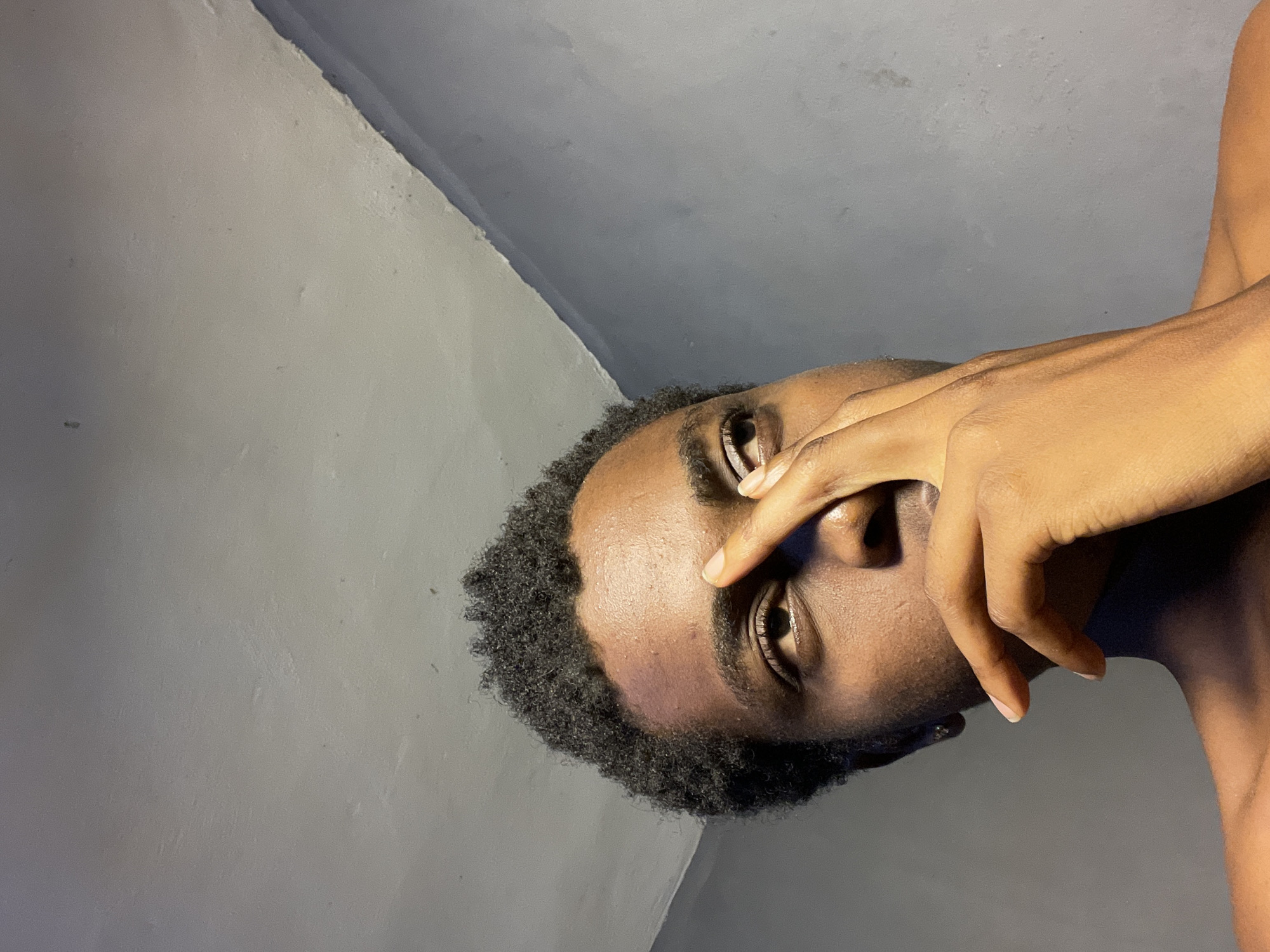 A close-up portrait of a Black man with short, curly hair, positioned at an unusual angle. The man's face is partially obscured by his hand, which is raised to his face. The background consists of gray walls. The image lacks context to confirm a location. The claim that this photograph was taken in Nkpor, Nigeria cannot be confirmed.