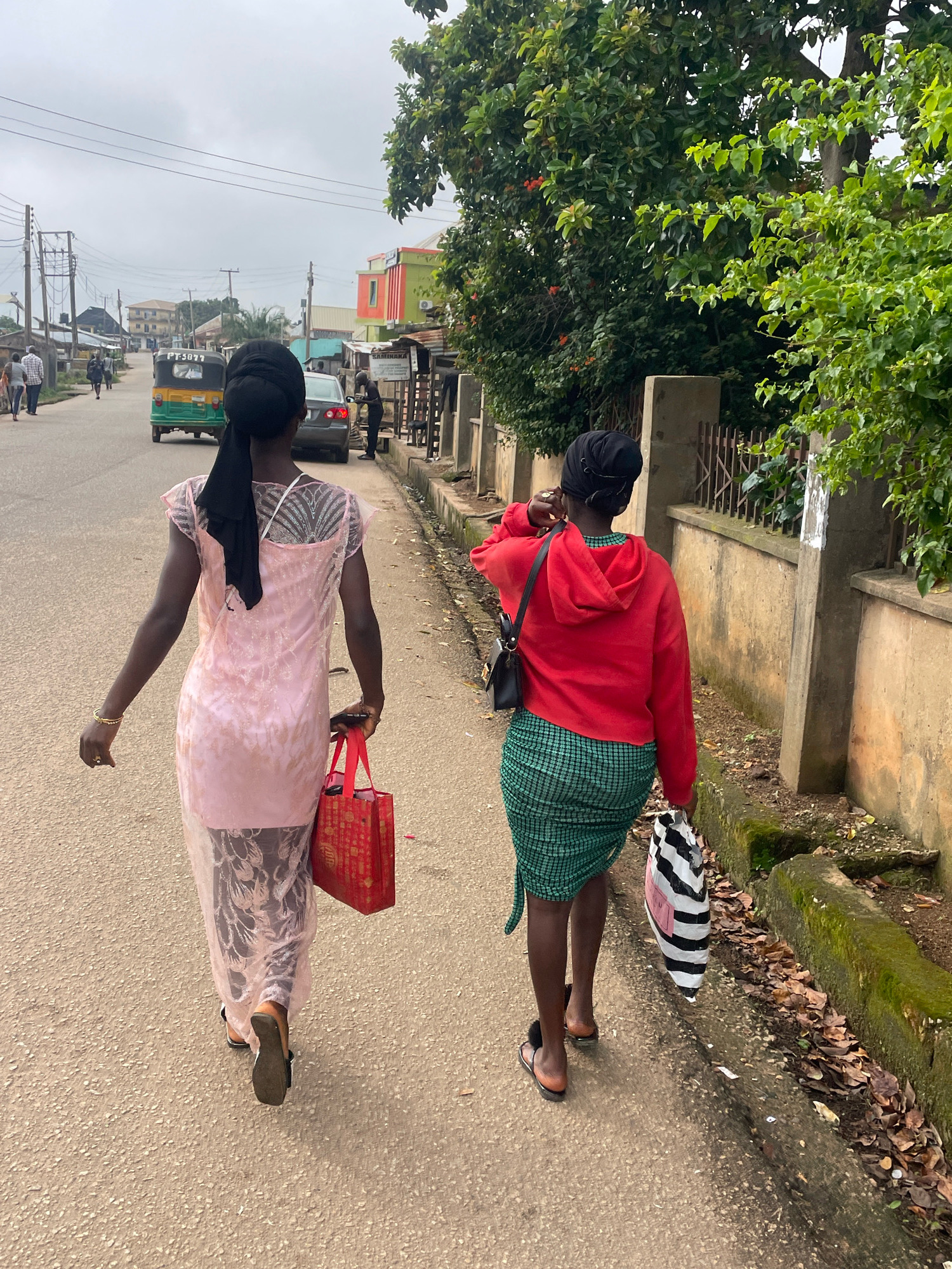 Two young women are walking away from the camera on a paved street in the city of Jos, Nigeria. The woman on the left is wearing a sheer pink dress over dark leggings and a black headscarf. She is carrying a red shopping bag in her right hand. The woman on the right is wearing a red hooded sweatshirt and a green and black checkered skirt. She has a black purse on her left shoulder and is carrying a striped shopping bag in her right hand. They are walking along the side of the road, which is lined with a low wall and overgrown vegetation. In the background, there are buildings, a tuk-tuk, and other pedestrians. The sky is overcast.