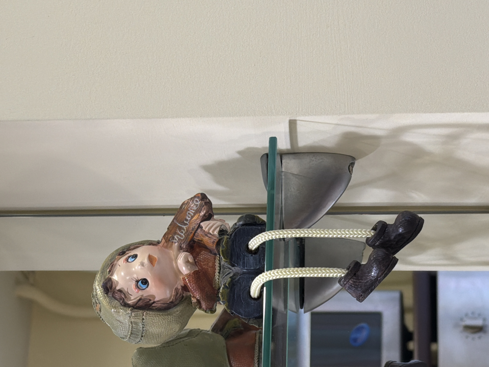A doll with blond hair, blue eyes, and wearing a gray-green hat and brown clothes is positioned facing the viewer but upside down. The doll is attached to a glass shelf by two strands of white cord that wrap around the doll's legs and attach to metal supports on the shelf. The background is mostly a neutral, matte white with some reflections from nearby objects on the glass shelf. The location of the image is unknown.