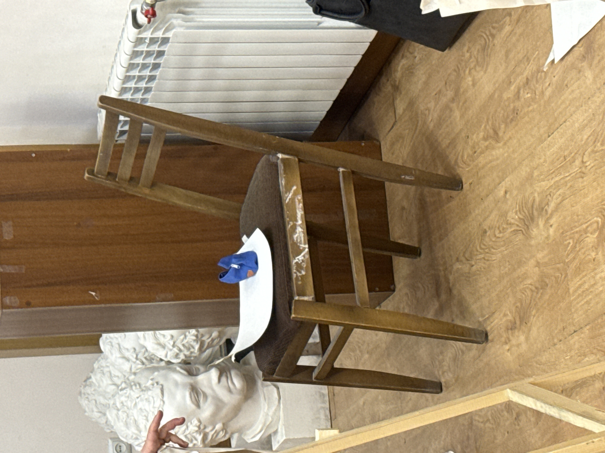 The image shows a room with a variety of objects. A white plaster bust sits near the bottom of the frame. Behind it is a chair with a blue object and white paper on its seat. Behind the chair, there is a wooden cabinet, and above that, a radiator. The room has a wooden floor, and the presence of a sculpture suggests the location could be an art studio or similar setting, potentially in Yerevan, Armenia. A hand is visible near the bottom left corner.