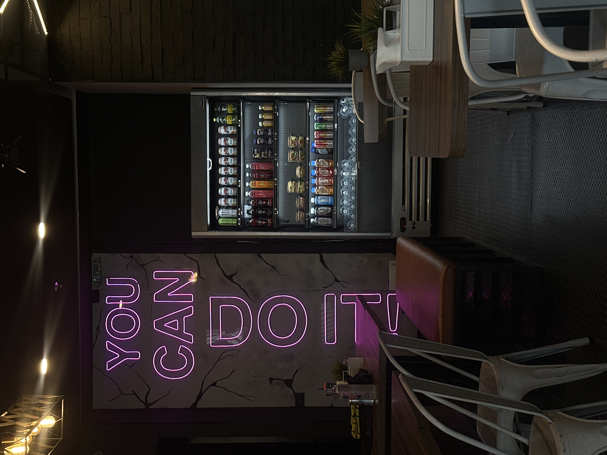 The image shows the interior of what appears to be a café or eatery. A vending machine stocked with beverages and snacks is visible against a dark wall, next to which a neon sign displays the phrase "YOU CAN DO IT!". The floor is dark and textured, possibly a rubberized surface. There are tables and chairs, with the chairs having white frames and stacked at the tables. The image is taken in a low-light environment.
There is no information about the city of San Bernardino Tlaxcalancingo, Mexico, in the image.