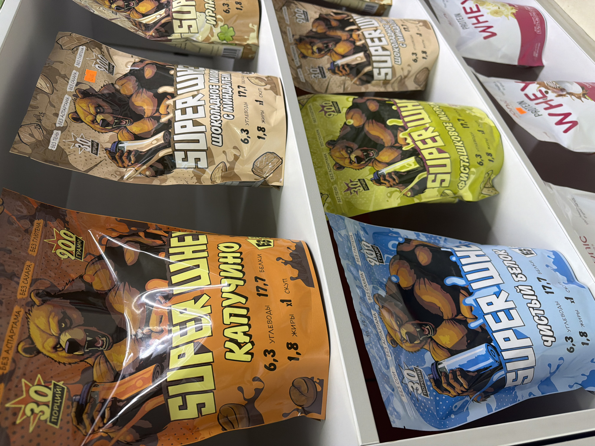 The image shows a retail shelf display of protein powder bags in a store located in Chaikovsky, Russia. The bags feature prominent graphics of muscular bears, a common marketing theme for sports nutrition products. Several flavors are visible, including "Cappuccino," "Chocolate Milk with Almonds," "Pistachio Mousse," and "Pure Whey." Nutritional information such as carbohydrates, protein, and fat content is also displayed on the packaging. The products are arranged on white shelves, suggesting a clean and organized retail environment. The lighting appears to be artificial, consistent with an indoor commercial space. The overall scene depicts a typical retail setting for health and fitness supplements.