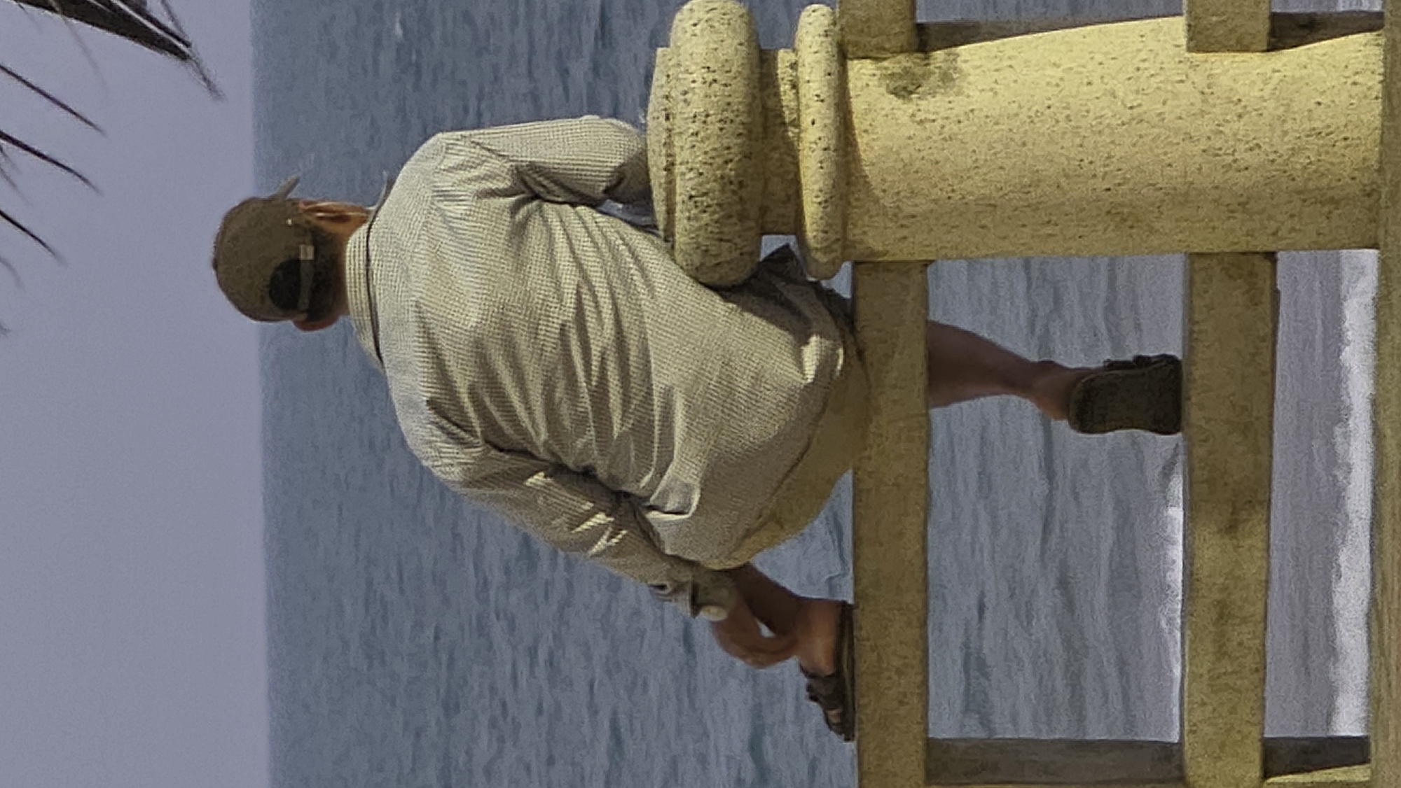 A man in a light gray button-down shirt and shorts sits on a yellow concrete railing overlooking the ocean. He wears a baseball cap and sandals. The railing is constructed with vertical posts and horizontal bars featuring rounded decorative elements. The ocean is in the background, along with an overcast sky. The left edge of the image shows parts of palm leaves. The photo was most likely taken in Nha Trang, Vietnam.