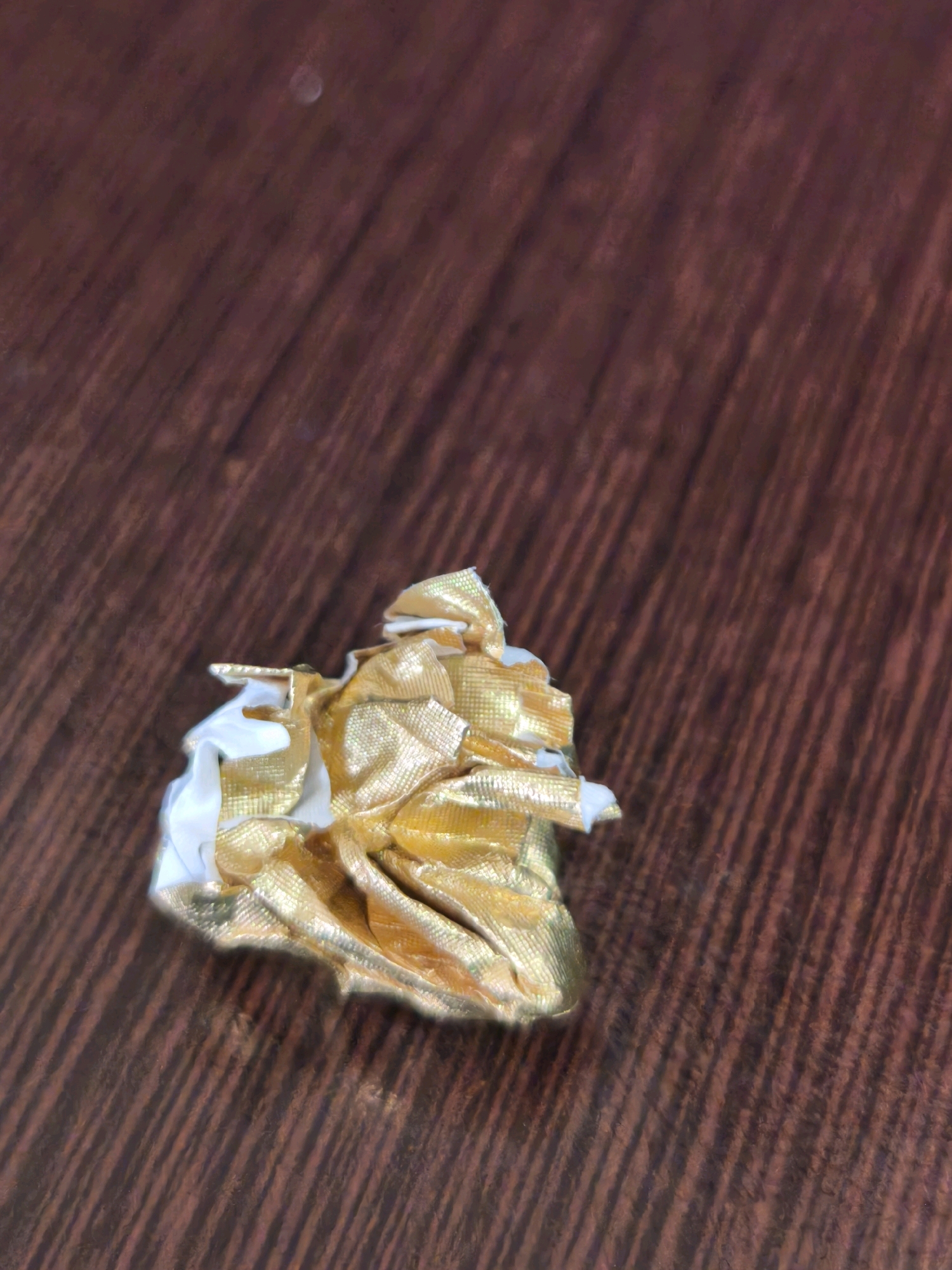 The image shows a crumpled piece of gold and white candy wrapper on a dark brown, wood-grained surface. The wrapper is small and appears to be made of metallic foil. The surface has a distinct pattern of diagonal lines, suggesting it is a table or countertop. The lighting is dim, casting subtle shadows. There is no indication of the city of Karachayevsk, Russia, or any people, actions, or interactions.