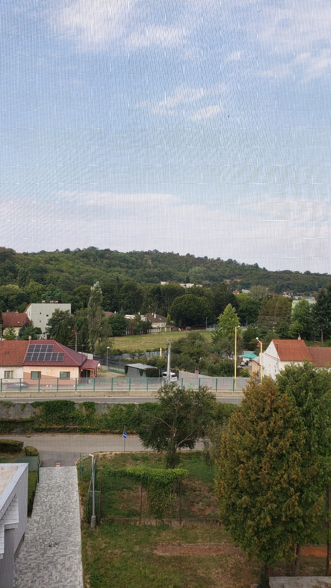 This image depicts a view from a window with a screen, overlooking a residential area with a forested hill in the background. The sky is partly cloudy. In the middle ground, there are several houses with red tile roofs and solar panels visible on one house. A road runs horizontally across the scene, bordered by a green fence and lush vegetation. A few trees are scattered throughout the landscape. In the foreground, a portion of a modern building with a grey facade and a paved walkway is visible. The scene appears to be in a suburban or semi-rural part of a city. The presence of a street sign and the architectural style of the houses suggest a European setting. Based on the provided context of Prešov, Slovakia, this view is likely from an apartment or house in a residential district of the city, possibly with a view towards the surrounding natural landscape or hills on its outskirts.