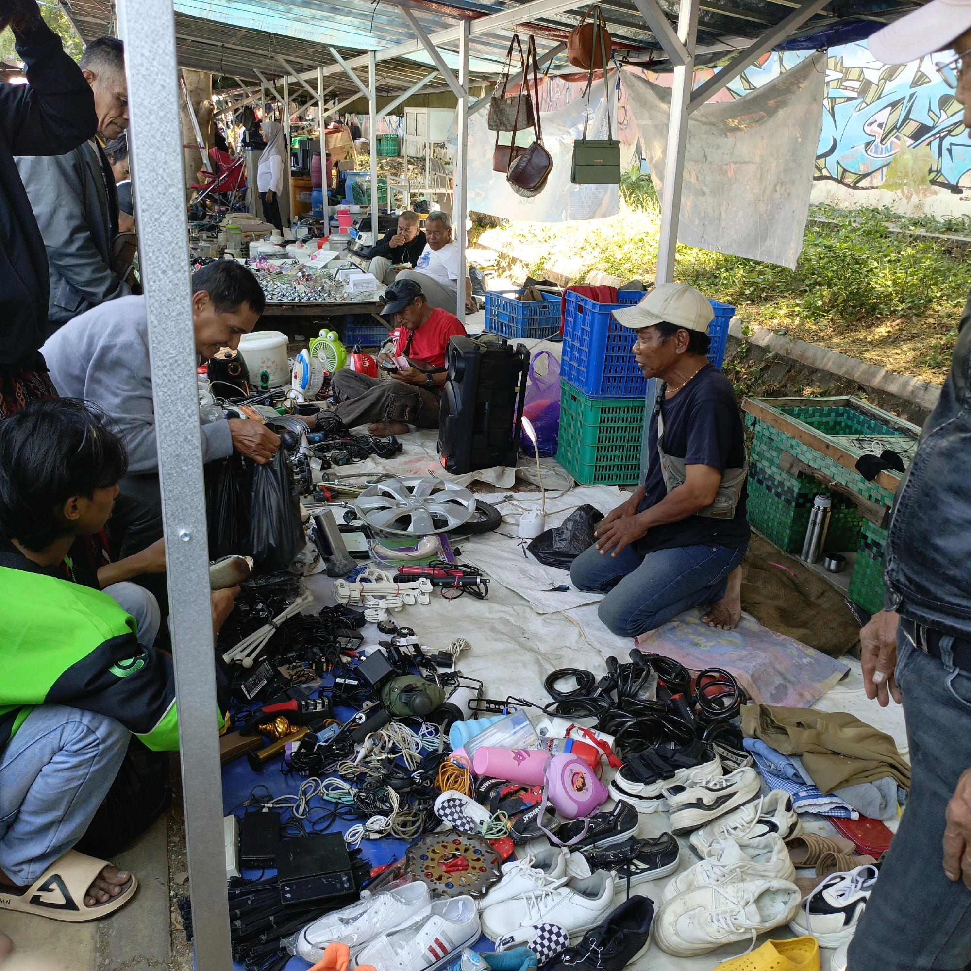 The image depicts an outdoor marketplace, likely a flea market or informal bazaar, in Ambarawa, Indonesia. A variety of second-hand goods are spread out on the ground on a white tarp, including electronics, cables, shoes, and car hubcaps. Several people are present, engaged in browsing or selling. One man, wearing a light grey sweatshirt and dark pants, is reaching into a black plastic bag. Another man, in a dark blue t-shirt and jeans, is kneeling and appears to be interacting with items on the tarp. A person wearing a bright green vest and blue jeans is also visible. In the background, there is a structure with a corrugated metal roof, draped with tarps and fabric. Graffiti is visible on a wall behind the market stalls. The overall scene suggests a bustling environment where used items are being traded.
