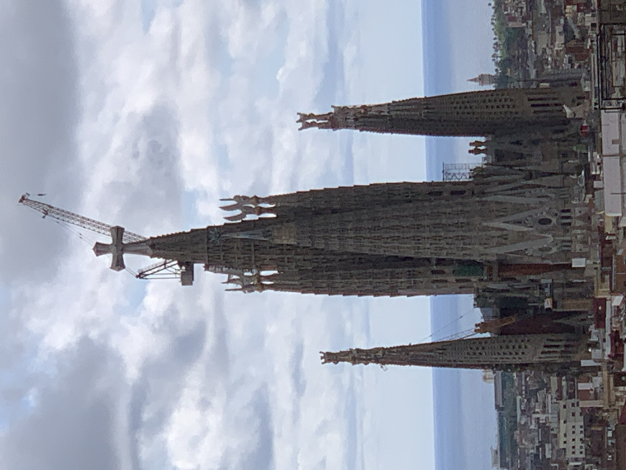 The image depicts the Sagrada Familia basilica in Barcelona, Spain, under construction. Several spires of the church are visible, one with a prominent construction crane attached. The surrounding cityscape of Barcelona extends to the sea in the background. The sky is cloudy. No people are directly visible in the image.