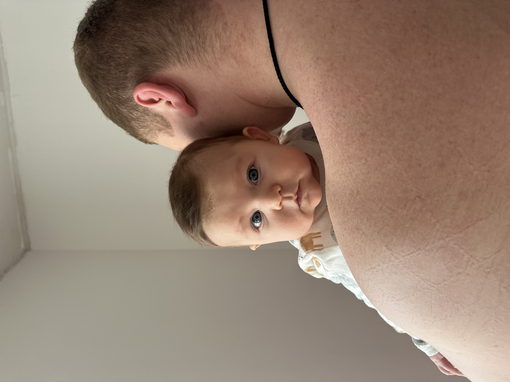 The image shows a baby with blue eyes being held. The baby is wearing a light-colored outfit with a pattern of brown animals. Behind the baby is a person with light skin, a short haircut, and a thin black necklace. The background appears to be a plain white wall. The location is not discernable in the image.