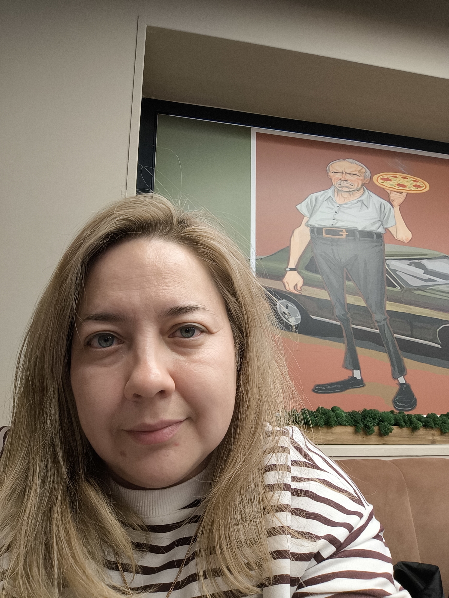 A woman is taking a selfie in what appears to be a restaurant. She has light blonde hair, blue eyes, and is wearing a white and brown striped long-sleeved shirt and a gold necklace. Behind her is a framed illustration of an older man holding a pizza, standing in front of a vintage car. The restaurant has a brown couch and a green, moss-like decoration. The city is Khimki, Russia.