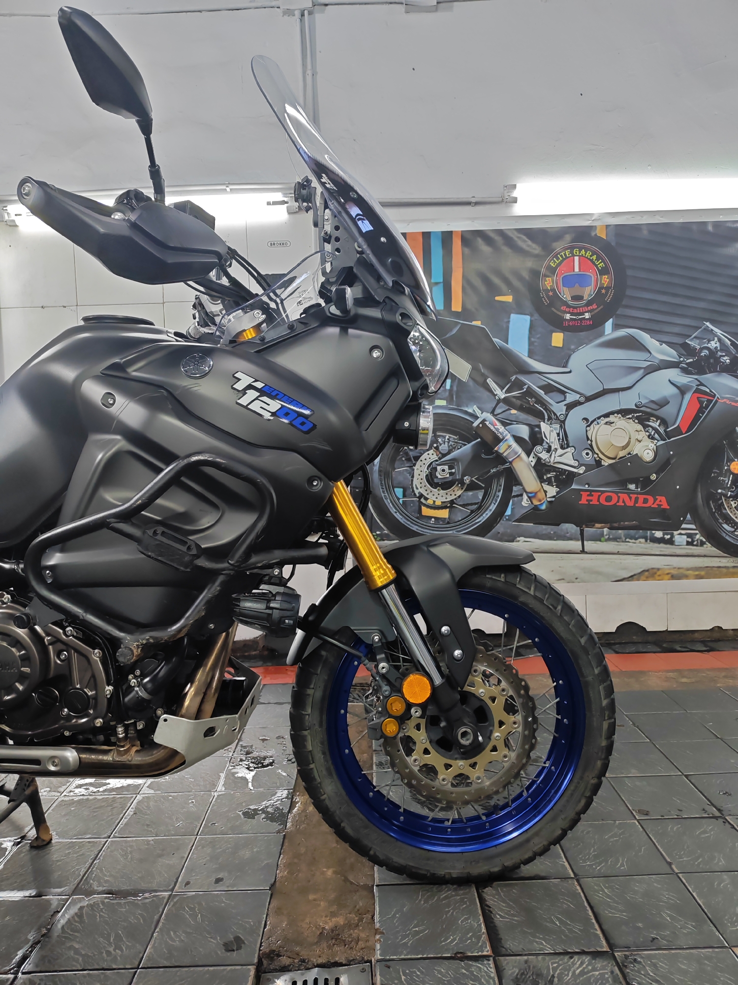 A close-up shot depicts the front portion of a matte black Yamaha Super Ténéré motorcycle. The motorcycle is parked indoors on a gray tiled floor. A blue rimmed front wheel with visible disc brakes is featured prominently. Behind the motorcycle, there is a large poster or wall graphic of a Honda motorcycle. The poster also displays the words "ELITE GARAJE" and "detailing." The lighting suggests an indoor setting with fluorescent lights, possibly a garage or workshop. The location is possibly in the city of Buenos Aires, Argentina based on the phone number "11-6912-2284" on the poster, which is a Buenos Aires area code.
