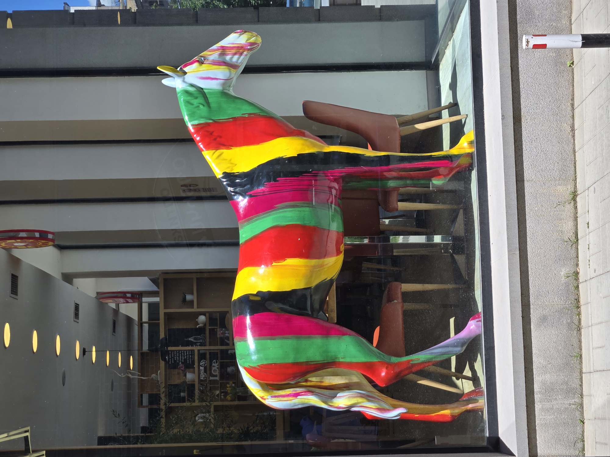 A vividly painted horse sculpture is displayed in a storefront window. The sculpture is predominantly white, with thick, vertical stripes of red, yellow, green, and black paint dripping down its body. The paint appears to be applied in a chaotic, artistic style, with some areas showing distinct drips and splatters. The horse is positioned to face right, with its head and neck slightly lowered.
Behind the sculpture, through the glass window, a cafe interior is visible. The cafe appears to have wooden shelves filled with various items, including what look like books and decorative objects. There are also tables and chairs inside, some of which are partially obscured by the horse sculpture. A line of yellow circular lights is visible on the ceiling of the cafe.
The storefront is situated on a city street. The exterior pavement is made of grey rectangular tiles, with some sparse green weeds growing between them. A white and black bollard with red stripes stands on the pavement to the right of the storefront. The sky above is a bright blue, indicating a sunny day. The overall scene captures a piece of public art displayed in an urban setting.