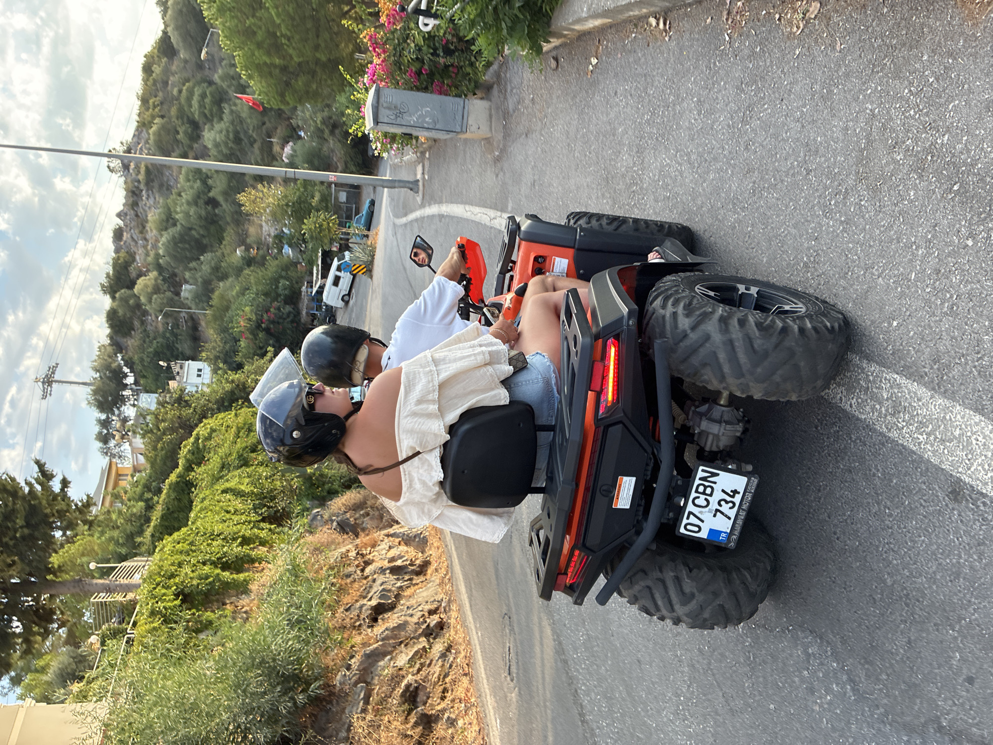 Two people are riding an orange ATV on a paved road in Alanya, Turkey. The ATV has a black seat and a license plate that reads "07 CBN 734". The person in the front, wearing a black helmet and sunglasses, is a woman with long brown hair. She is wearing a cream-colored off-the-shoulder top and denim shorts. The person behind her, wearing a white t-shirt and a black helmet, is likely a man. They are riding up a slight incline on a road lined with rocky terrain and lush green vegetation on the left, and a sidewalk and more vegetation on the right. In the background, there are buildings, trees, and a cloudy sky. The overall scene suggests a recreational activity in a scenic, hilly area of the city.