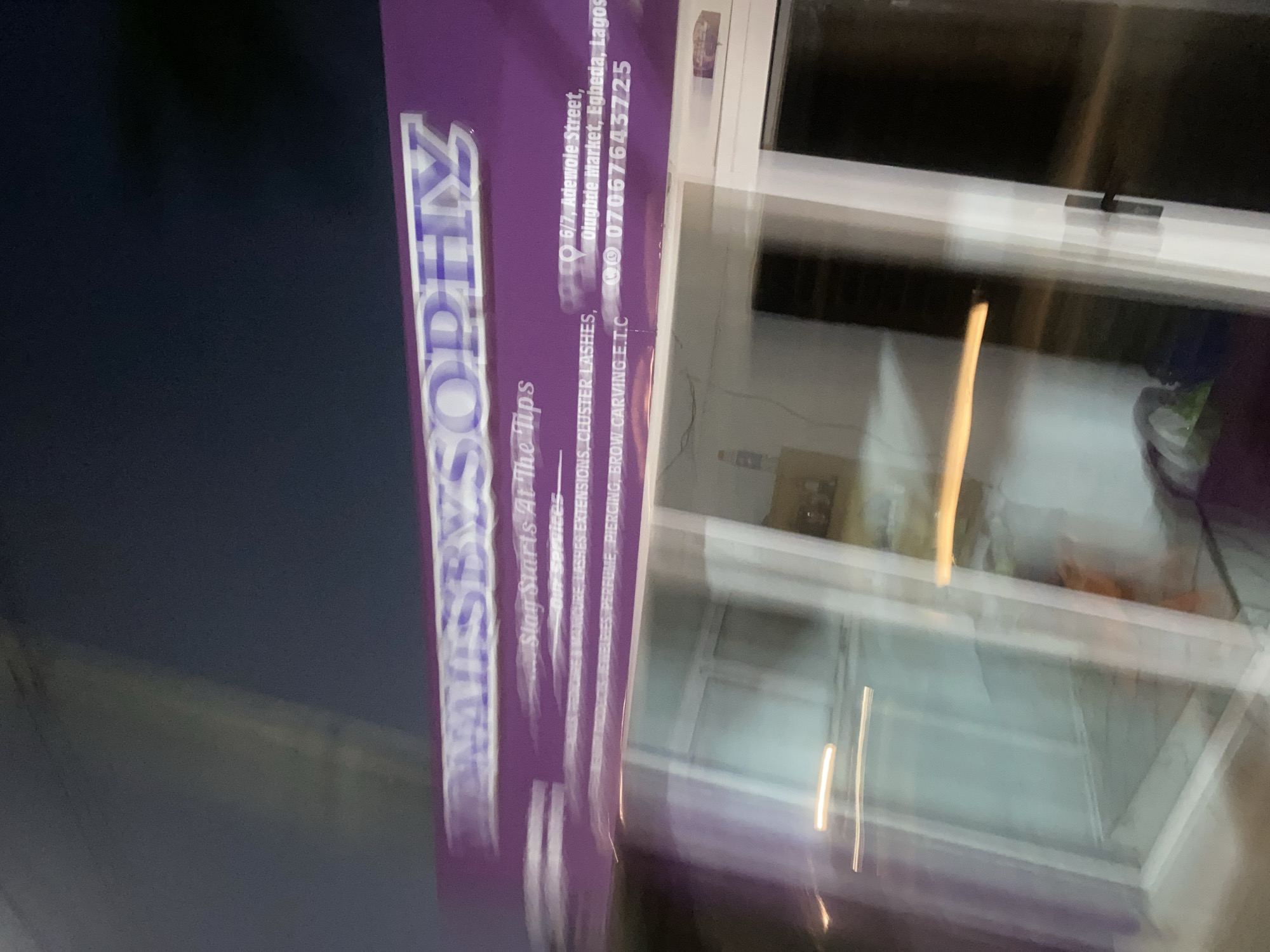A blurry, slightly out-of-focus image shows a sign for "Mistasy Sophyz", a business offering services like lashes extensions, cluster lashes, and piercing and brow carving, located at 6/7 Adewole Street, Olugbde Market, Egbeda, Lagos, Nigeria. The phone number provided is 07067643725. The sign is purple and white and is situated next to a building with window frames. Due to the blur, finer details are not discernible.