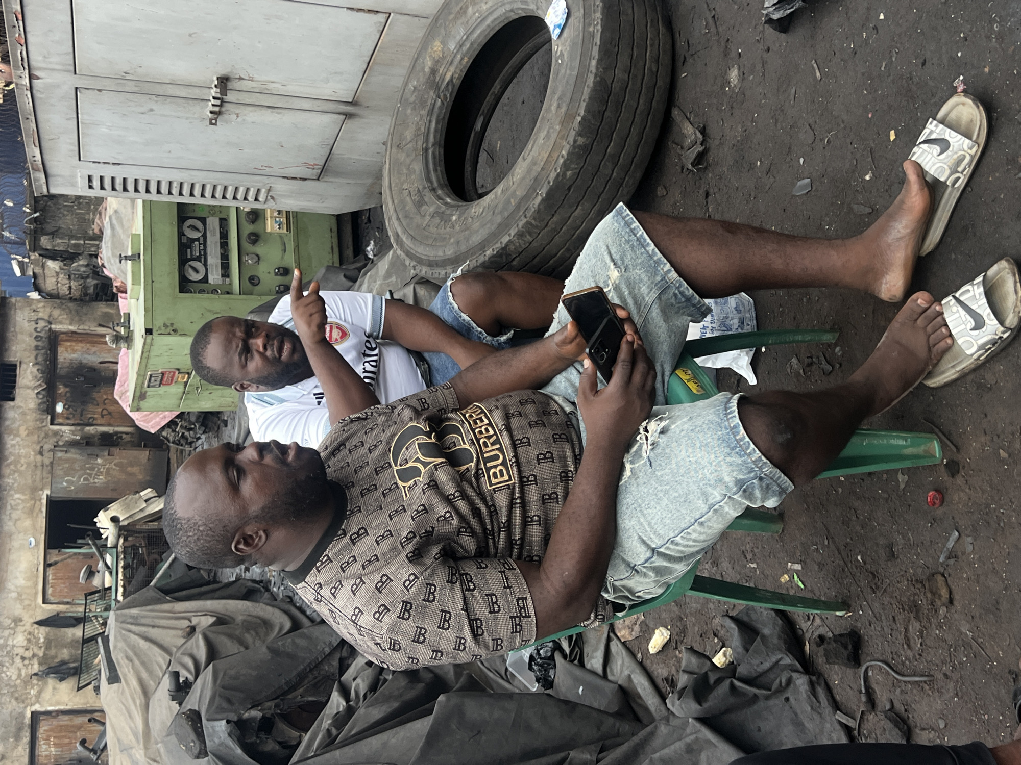 In Obosi, Nigeria, two men are seated outdoors amidst a cluttered industrial or workshop setting. The man on the left, wearing a white t-shirt with a red and white logo, is looking to the right and gesturing with his left hand. The man on the right, dressed in a patterned brown t-shirt and denim shorts, is holding a black smartphone in his right hand, looking down at it. A large tire is visible behind the man on the left, and a green generator or industrial machine is behind them. The ground is dirt and littered with debris. The overall scene suggests a candid moment, possibly a break in work. The lighting indicates it is daytime.