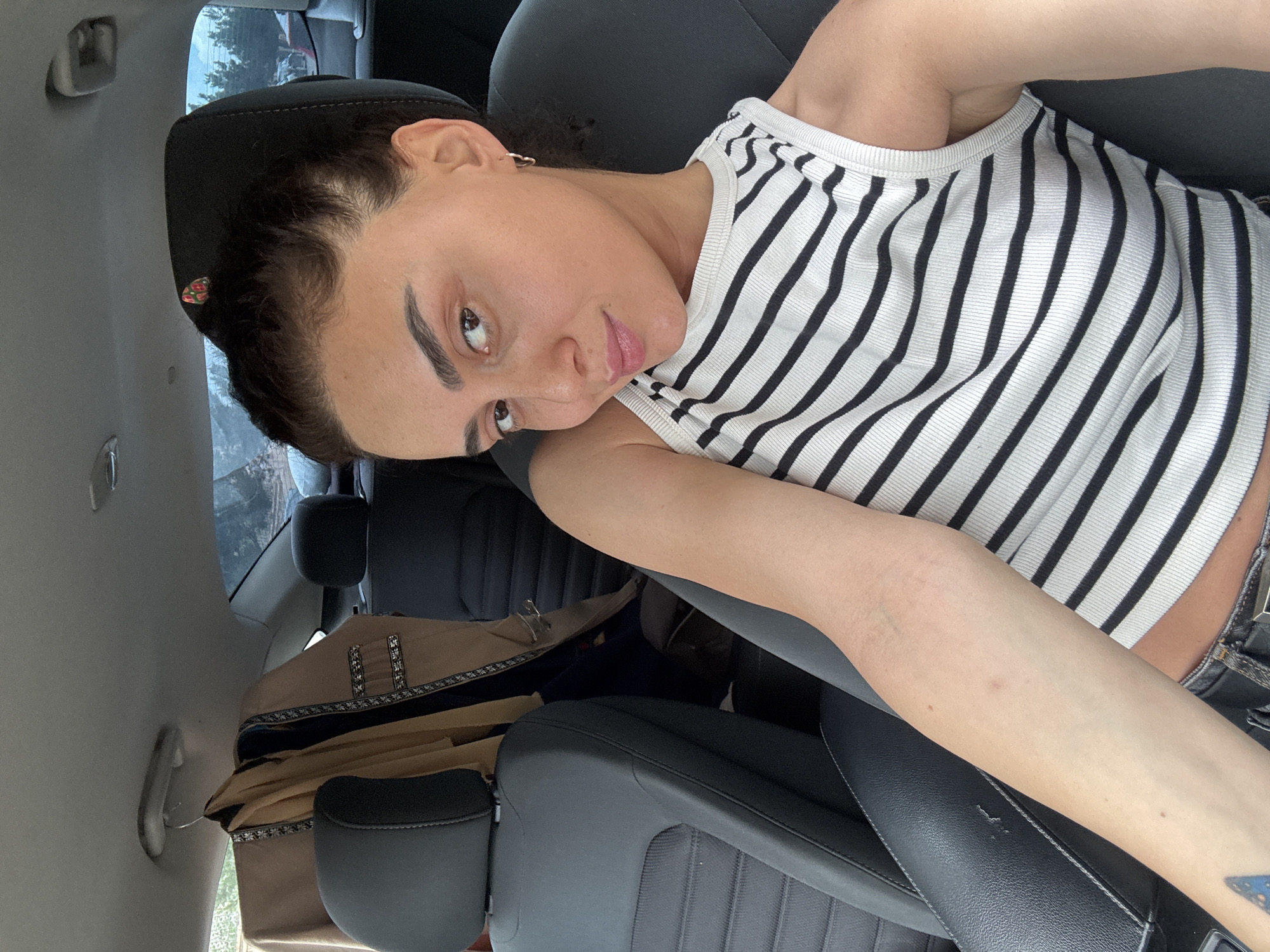 A young woman with dark hair, pulled back into a bun, sits in the passenger seat of a car. She is wearing a black and white striped tank top. Her left arm is resting on the seat beside her, and her right arm is bent at the elbow, with her hand near her face. A jacket or coat is hanging on a hanger from the ceiling of the car, behind the passenger seat. The interior of the car appears to be dark gray or black. Outside the car windows, trees and foliage are visible, suggesting a natural or suburban setting. The image does not contain any elements that would indicate it was taken in the city of Tbilisi, Georgia.