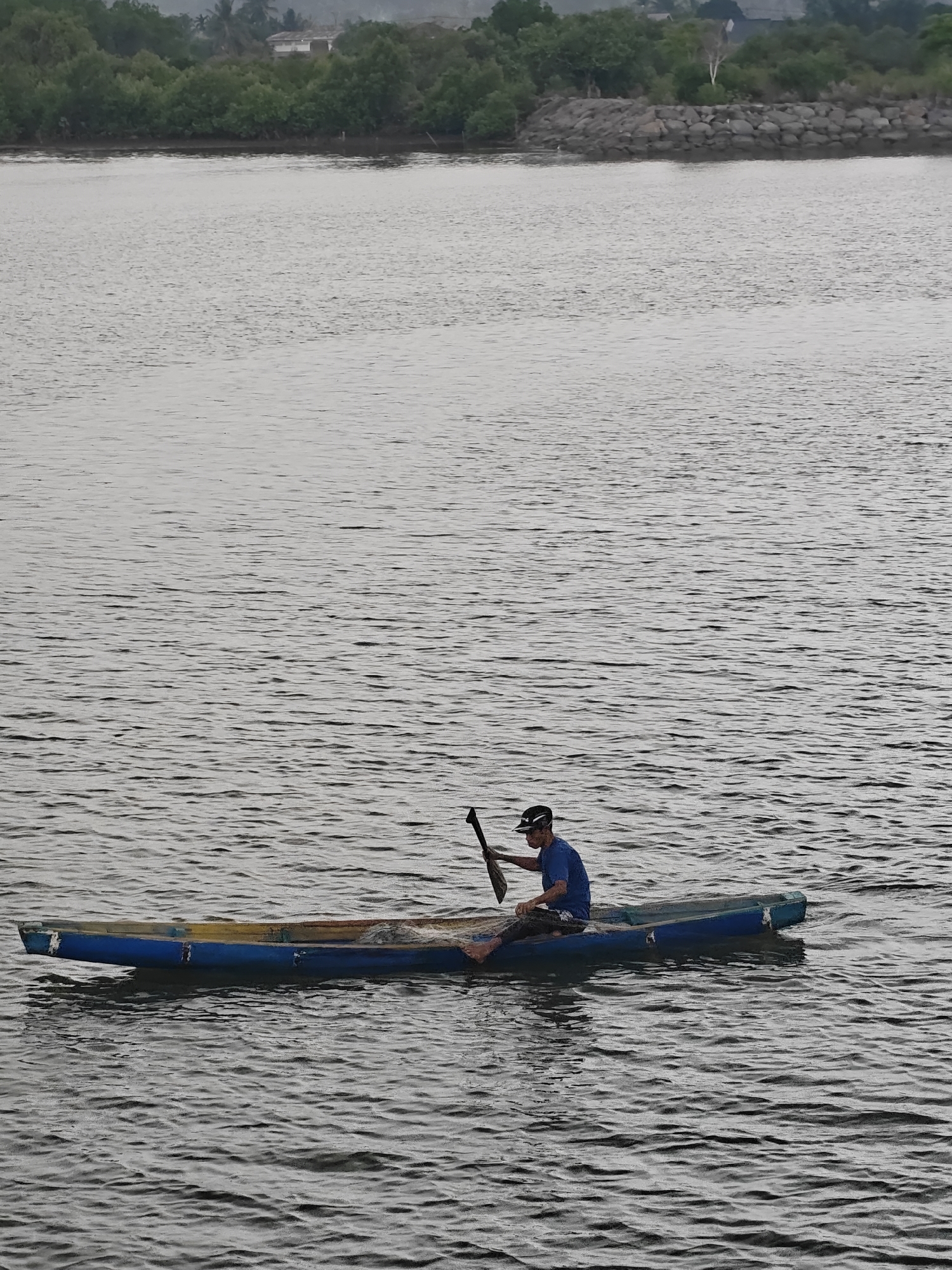 The photograph shows a man in a blue shirt and a hat paddling a blue and yellow boat on a body of water. The man is using a single paddle, positioned on his right side. The water is rippled, indicating a slight breeze or movement. In the background, there is a coastline with trees and what appears to be a stone barrier. Based on the geographical features, this photo was likely taken in Lhokseumawe, Indonesia.