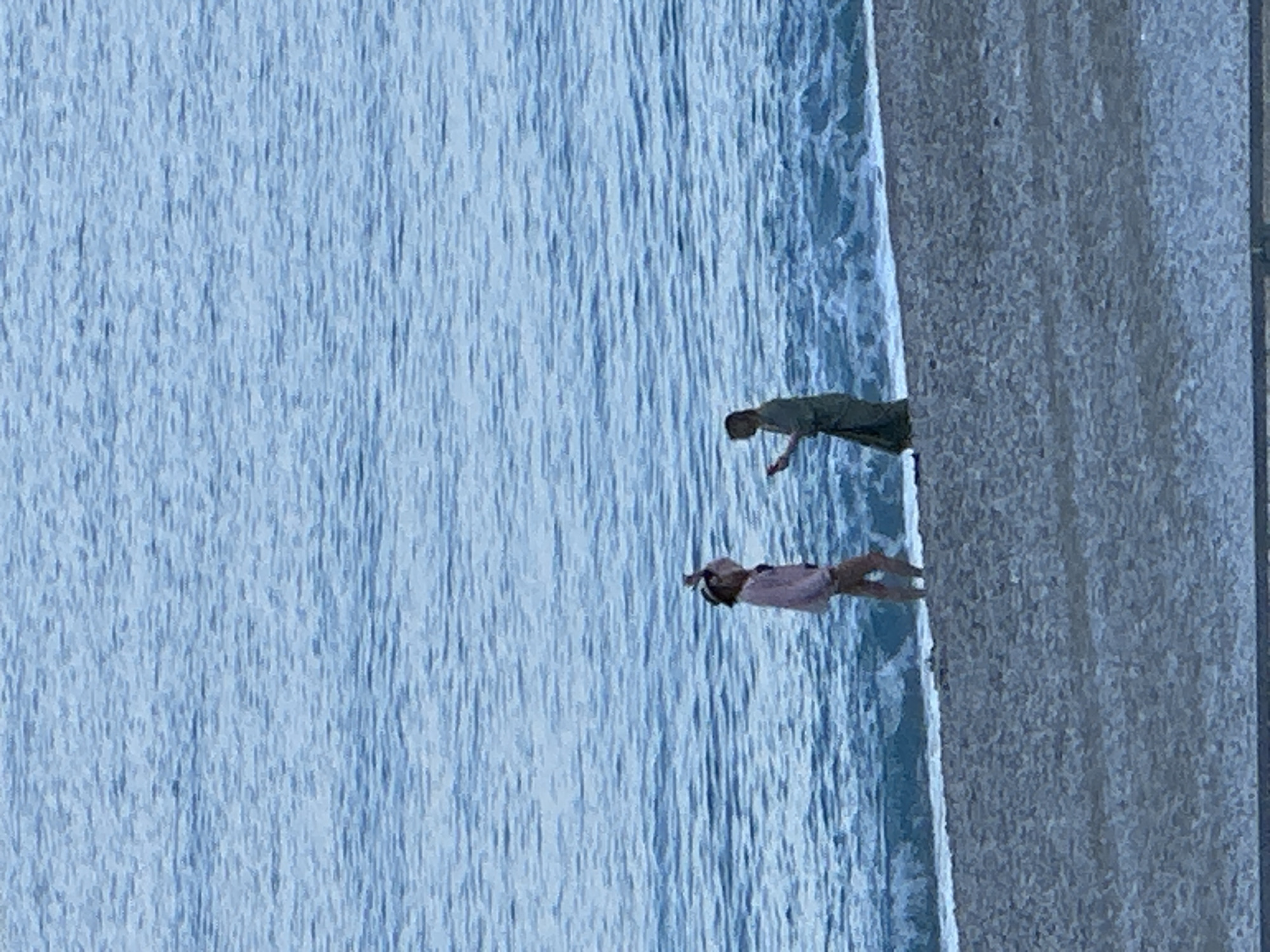 The image depicts an outdoor scene at a beach in Fethiye, Turkey, under diffused natural light. Two individuals stand on the sandy-pebbled shore where small waves meet the land. The calm, blue-grey sea extends into the background, showing ripples and gentle undulations.
The person on the left, positioned closer to the water's edge, wears a light-colored (possibly pink or beige) long-sleeved top and light-colored bottoms. They are seen with both hands raised, holding a small, light-colored object, possibly a smartphone, near their head, potentially taking a photograph, recording video, or adjusting something.
The person on the right, standing slightly further back from the water, wears a dark, possibly olive green or greyish-green, long dress or robe. This individual faces the first person and holds a dark-colored object, consistent with a smartphone, in their left hand. Their posture suggests they are either interacting with the device or engaging with the first person. The two individuals are in close proximity, indicating an interaction.