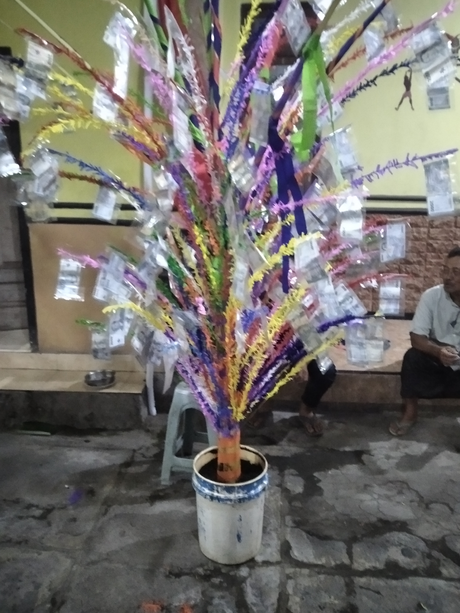 The image shows a decorative tree with colorful streamers and what appear to be bills attached to its branches, planted in a white bucket on a stone floor. The background reveals a building with yellow and brown walls. A person in a light-colored shirt is seated to the right, and another person's legs are visible near the tree. A light-colored stool is placed behind the tree. The scene is illuminated by artificial light, suggesting it is nighttime or an indoor setting. The overall atmosphere is festive or ceremonial. The location is identified as Rambipuji, Indonesia.
