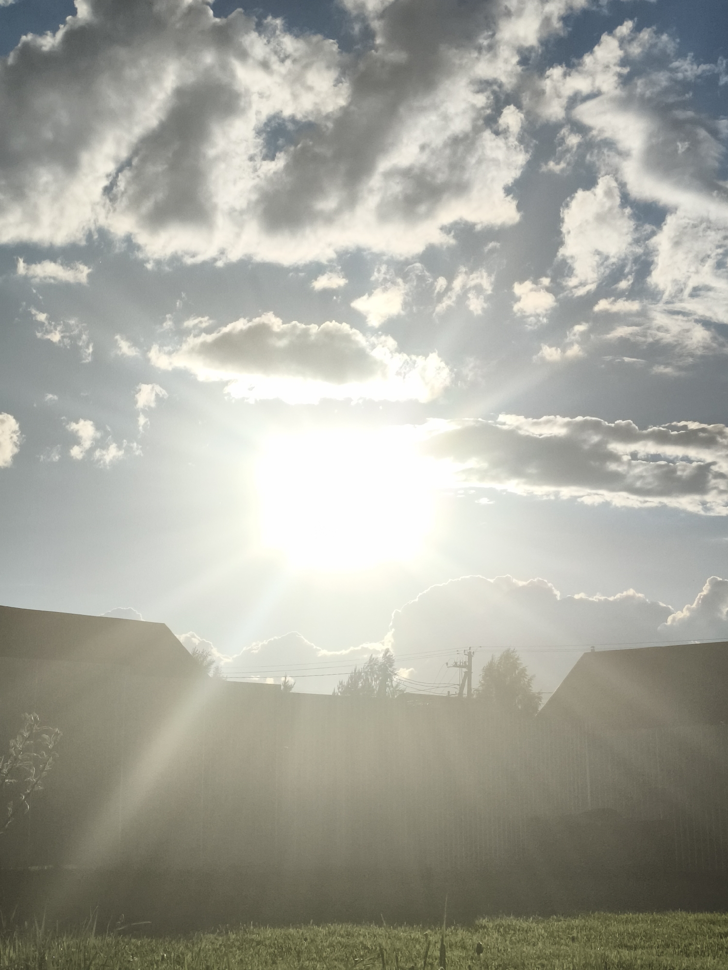 The image shows a sunlit scene in what appears to be a rural or suburban area. The sky is filled with clouds, some of which are illuminated by the bright sun, which is positioned high in the sky. The sun's rays are visible, casting a strong light downwards. In the foreground, there is green grass. Further back, there is a fence and the roofs of buildings or houses are visible. Power lines and trees are visible in the midground, suggesting the location is populated. The image appears to be taken in an outdoor environment during daylight. The provided location, Русский Вожой, Russia, suggests this is a scene from that specific area, however, there is no way to confirm it in the photo. There are no people present in the image.